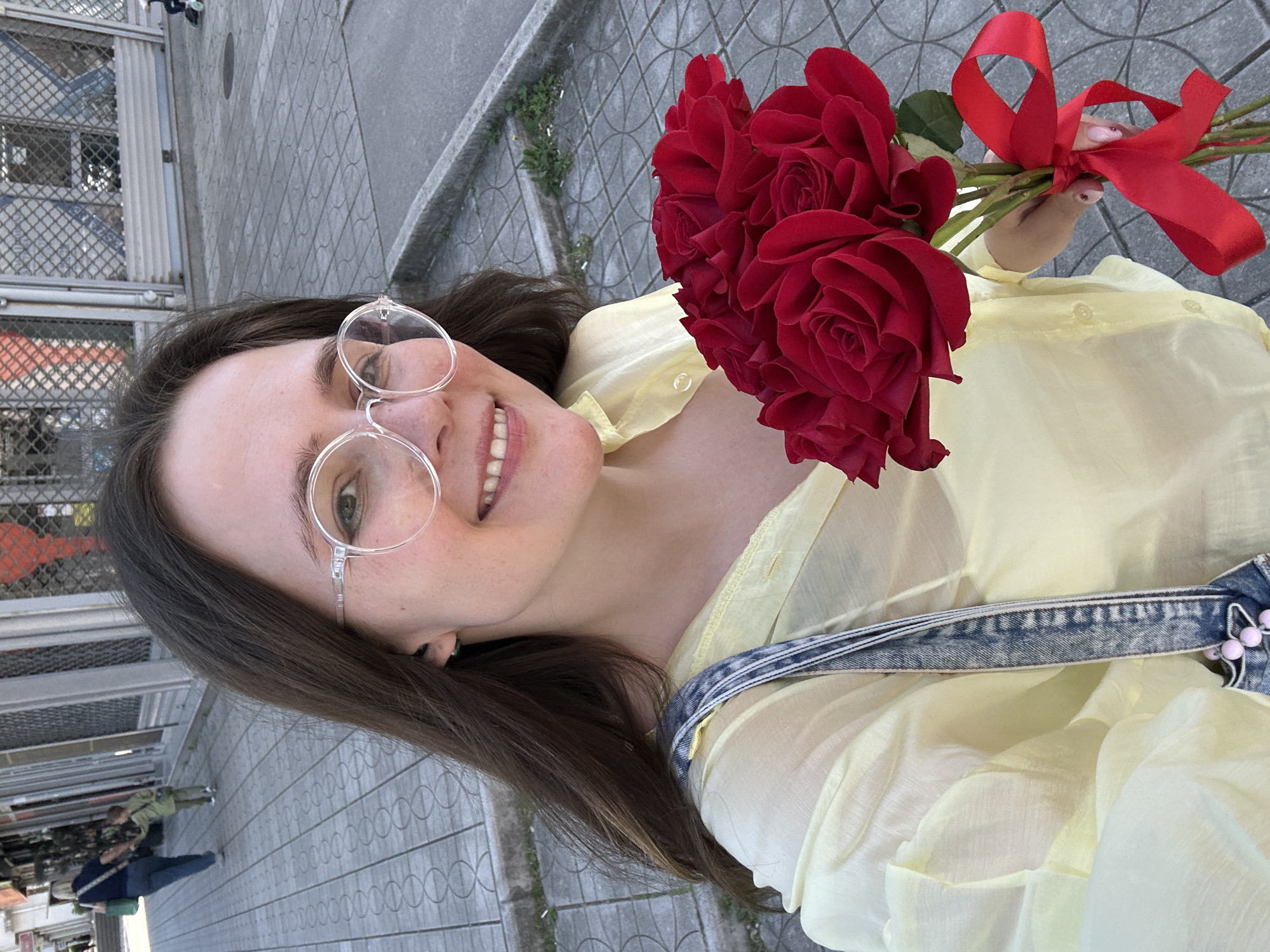 A young woman with long brown hair and wearing clear-framed glasses smiles while holding a bouquet of red roses tied with a red ribbon. She is wearing a light yellow, button-down shirt and denim overalls. The photo is taken at an angle outdoors, with a sidewalk and a fence visible in the background, suggesting an urban setting in Šušanj, Montenegro. Other people are seen further in the background.