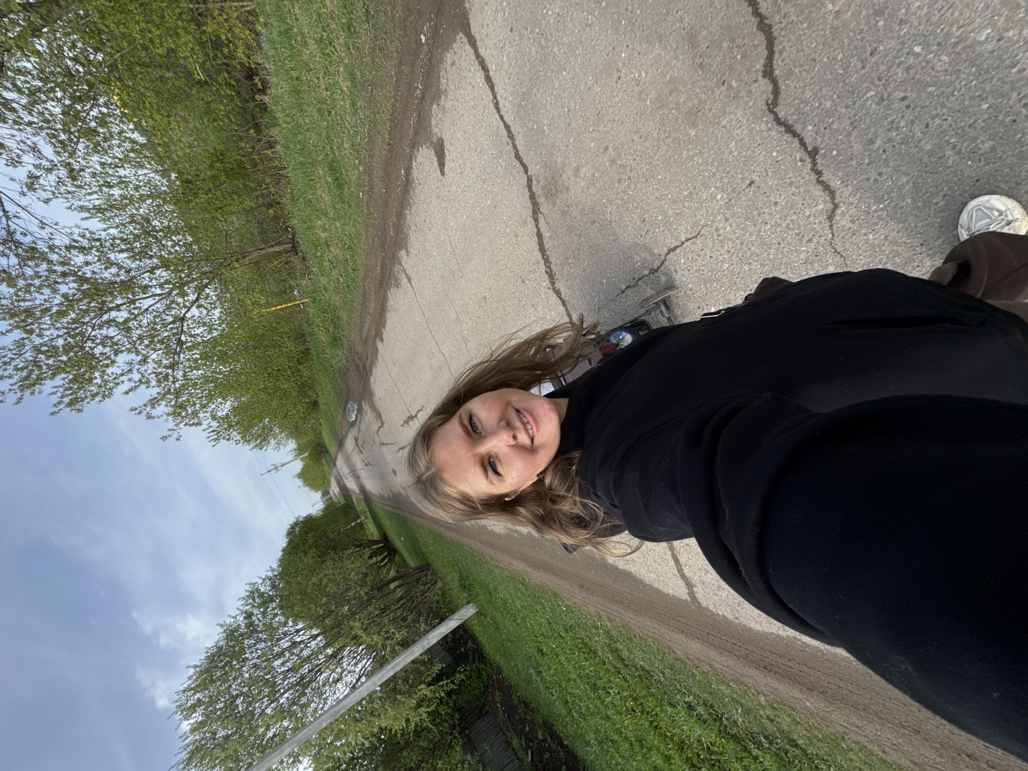 A selfie captures a person on a cracked pavement road bordered by grass and trees, suggestive of Горный, Russia. The person is wearing a black jacket. A skateboard is beneath their feet. The sky overhead is partly cloudy. A utility pole is visible along the roadside.