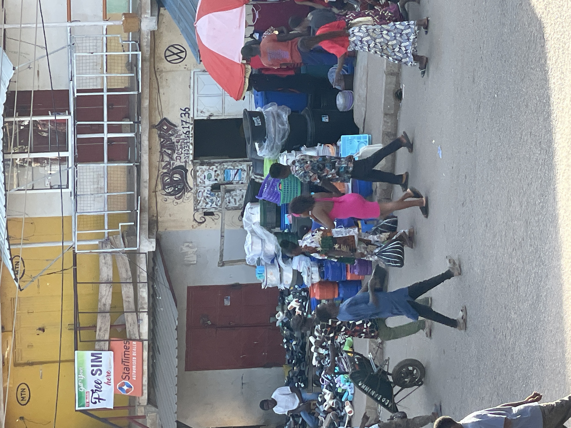 The image captures a bustling street scene in Bukuru, Nigeria. Various vendors are displaying their merchandise, including a large assortment of flip-flops and sandals on the lower right side, stacks of colorful plastic buckets, and other household items. Several people are walking through the scene, some of whom appear to be shopping.
Signage is visible on the left side of the image, including MTN and StarTimes banners advertising free SIM cards and authorized dealer services. Graffiti and phone numbers are visible on the buildings in the background. A red and white umbrella provides shade over one of the vendor's displays.