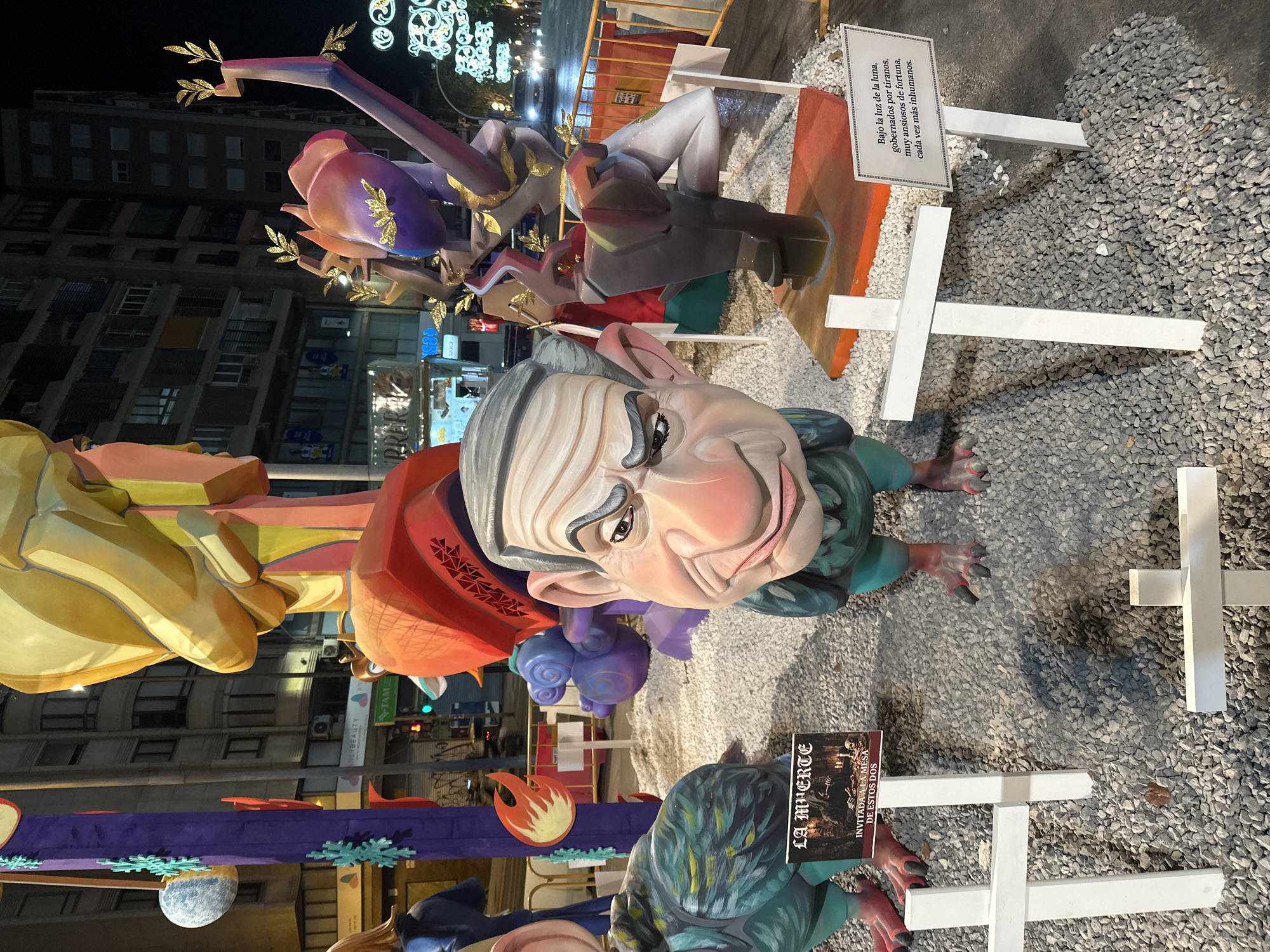 The image depicts a nighttime outdoor scene in Alicante, Spain, likely during the Las Fallas festival. The foreground is dominated by large, colorful figures characteristic of Fallas sculptures. In the center, a caricature of a man with exaggerated features and a grim expression stands with arms down. To the left and right of this central figure are large, stylized bird-like sculptures. Several white crosses are planted in the gravel surrounding the figures, suggesting a theme of death or remembrance.
Two signs are visible. One, placed near the left bird-like figure, has the title "LA MUERTE" (Death) and an image of skeletons around a table. Below this, it reads "INVITADA A LA MESA DE ESTOS DOS" (Invited to the table of these two). The second sign, near a cross on the right, features text in Spanish that translates to "Under the moonlight, governed by tyrants, very eager for fortune, each time more inhuman."
In the background, buildings and streetlights are visible, indicating an urban setting. Cars are present on the street, and traffic signals are illuminated. The overall atmosphere is festive, with the brightly lit sculptures contrasting with the nighttime environment. The presence of Fallas sculptures and the specific thematic elements point to this being a public installation during a festival in Alicante.