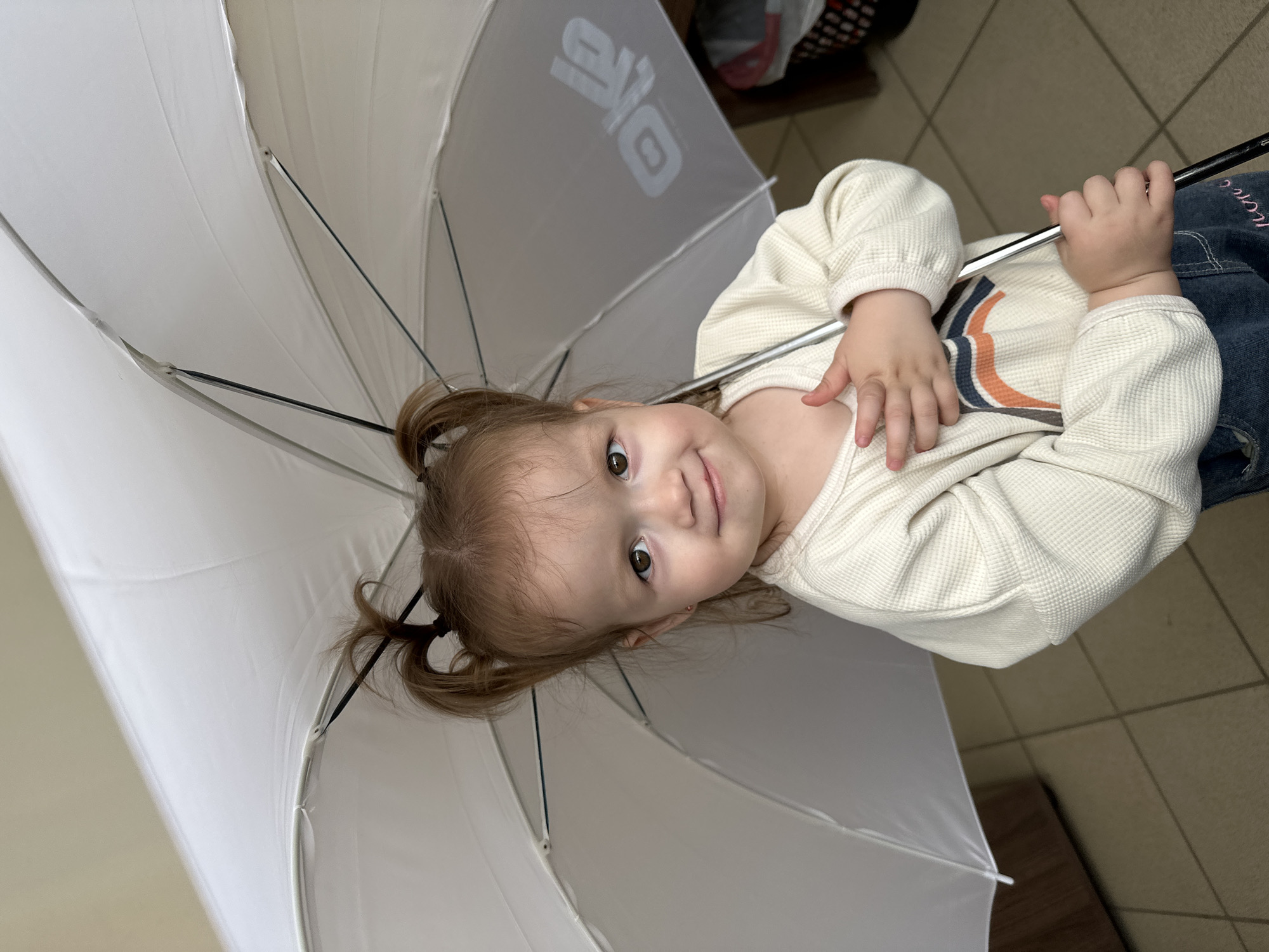 A young child, likely a girl, stands holding a large, white umbrella over her head. She has light brown hair pulled into two pigtails that stick out. Her eyes are brown, and she has a slight smile on her face. She is wearing a light-colored, waffle-knit long-sleeved shirt with a colorful stripe design across the chest and blue jeans. Her hands are clasped together, one resting on her chest and the other holding the handle of the umbrella. The background shows a tiled floor and what appears to be the corner of a wooden cabinet or desk. The umbrella has a logo printed on it, which is partially visible. There is no identifiable information to suggest this image was taken in Yekaterinburg, Russia.