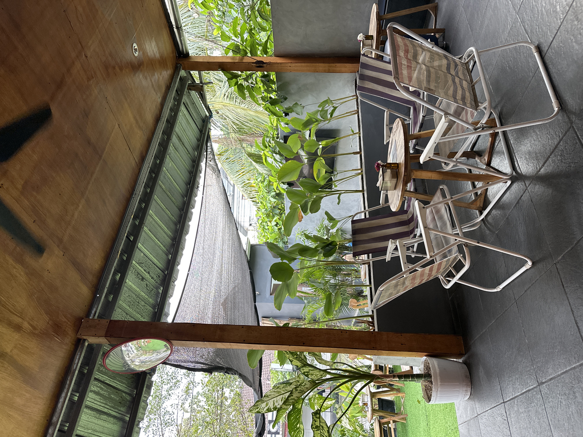 This is an outdoor seating area at a cafe in Kuta Selatan, Indonesia. The scene is covered by a wooden roof supported by dark gray pillars. A black mesh shade hangs from the roof structure. The seating area features several outdoor chairs with striped fabric and small wooden tables. One table has a tissue dispenser and two small bottles, one with a single flower. Another table in the background appears to have a drink and a small bottle with a flower.
To the left of the seating area is a potted plant with variegated green and white leaves. Beyond this, a patch of artificial green turf is visible with some wooden tables and chairs. In the background, there are several large green plants and a gray wall.
A convex mirror is mounted on a wooden post near the entrance to the seating area. The floor is tiled with dark gray square tiles. The overall impression is a relaxed, shaded outdoor dining space with ample greenery.