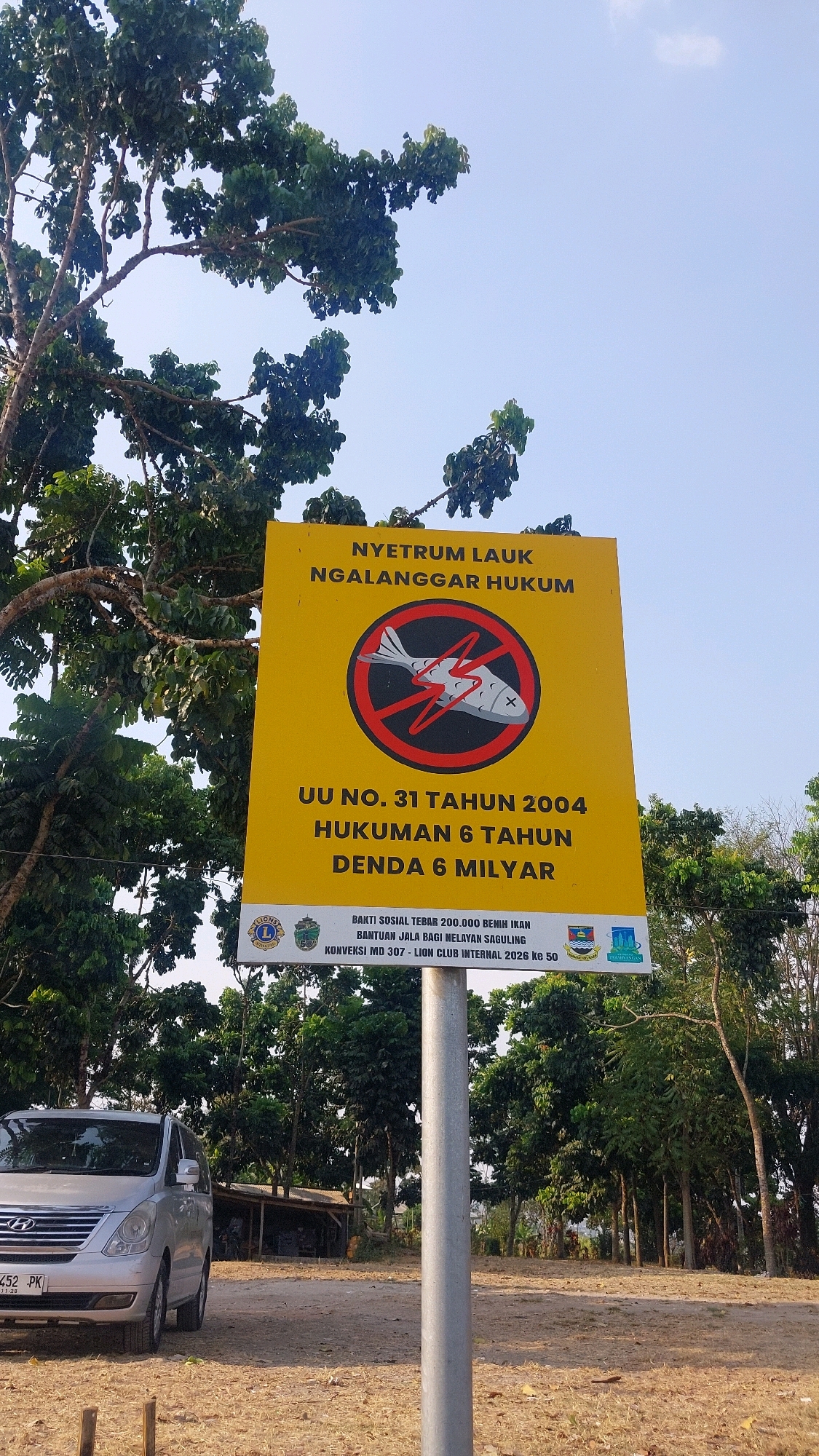The image shows a yellow sign with black text and a red circular prohibition symbol containing a fish with an 'X' through it, indicating a ban on electrofishing. The sign's text, in Indonesian, states "NYETRUM LAUK NGALANGGAR HUKUM" (Electrofishing Violates the Law), "UU NO. 31 TAHUN 2004" (Law No. 31 of 2004), "HUKUMAN 6 TAHUN" (6 Years Sentence), and "DENDA 6 MILIYAR" (Fine of 6 Billion). Below this are smaller texts mentioning "BAKTI SOSIAL TEBAR 200.000 BENIH IKAN" (Social Service Distribute 200,000 Fish Seeds), "BANTUAN JALA BAGI NELAYAN SAGULING" (Net Assistance for Saguling Fishermen), and "KONVERSI MD 307 - LION CLUB INTERNAL 2026 KE 50". The sign also features logos of Lions Club International and what appears to be a local government emblem. The sign is mounted on a metal pole.
The background consists of lush green trees and a clear blue sky with a few wispy clouds. A silver minivan is parked on the left side of the image, with its license plate visible as "B 1452 PK". A simple structure, possibly a shed or small building, is visible behind the minivan. The ground is covered with dry grass and dirt. The setting appears to be a rural or semi-rural area, consistent with the location described as Bojonghaleuang, Indonesia. There are no people visible in the image.