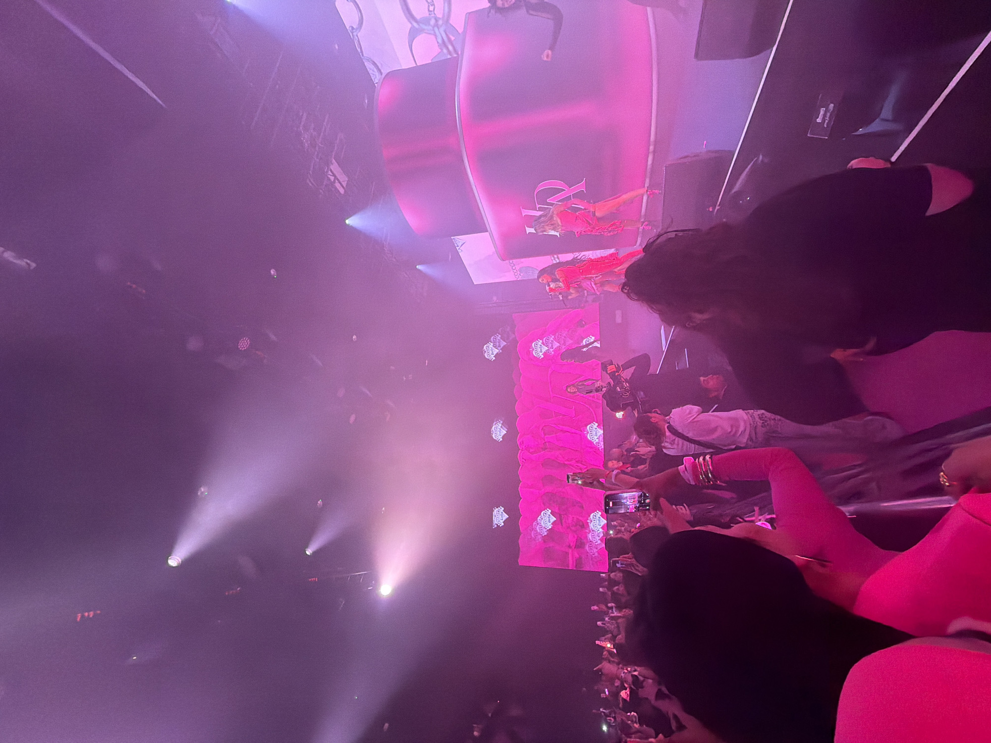 The image shows a live music performance viewed from the audience, bathed in pink and purple stage lighting. Figures on stage are visible on large screens, along with illuminated graphics. In the foreground, audience members are holding up phones to record the show. The crowd is dense and tightly packed. The location is likely a concert venue in London, United Kingdom, given the style of the show and the presence of stage lighting and large screens.