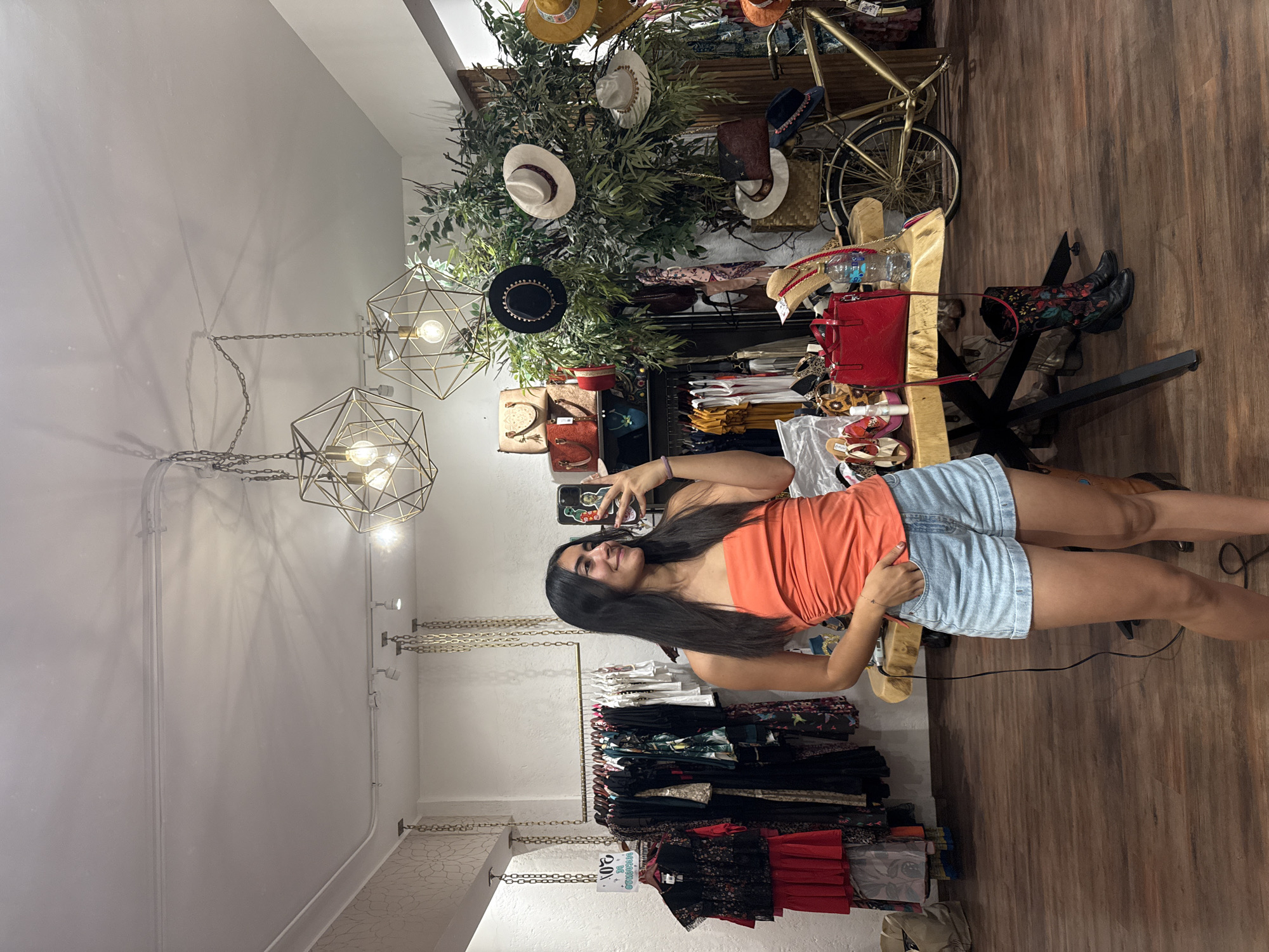 The photograph shows a woman taking a selfie in a clothing store in Domingo Arenas, Mexico. She is wearing an orange top and denim shorts. The store features clothing racks filled with garments, decorative hanging lights, and a display arrangement that includes hats, handbags, a bicycle, and boots. The floor is wooden.