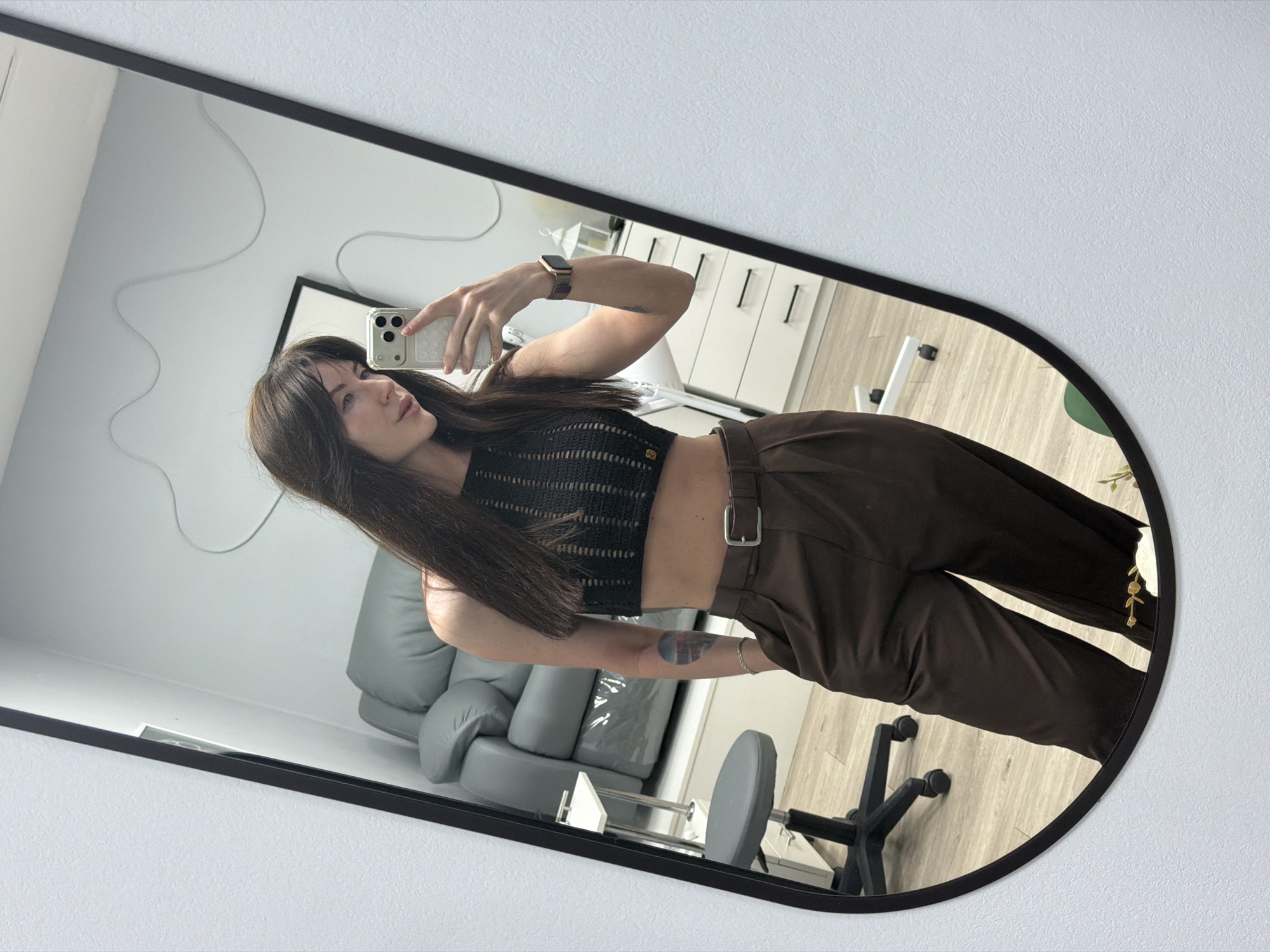 A woman with long, dark hair is taking a mirror selfie. She is wearing a dark-colored, cropped top and dark-colored, high-waisted pants with a belt. She is holding a white smartphone. The mirror is oval-shaped with a black frame. Reflected in the mirror is part of a room with a light-colored sofa, a chair, and light-colored walls. The location is possibly in Perm, Russia.