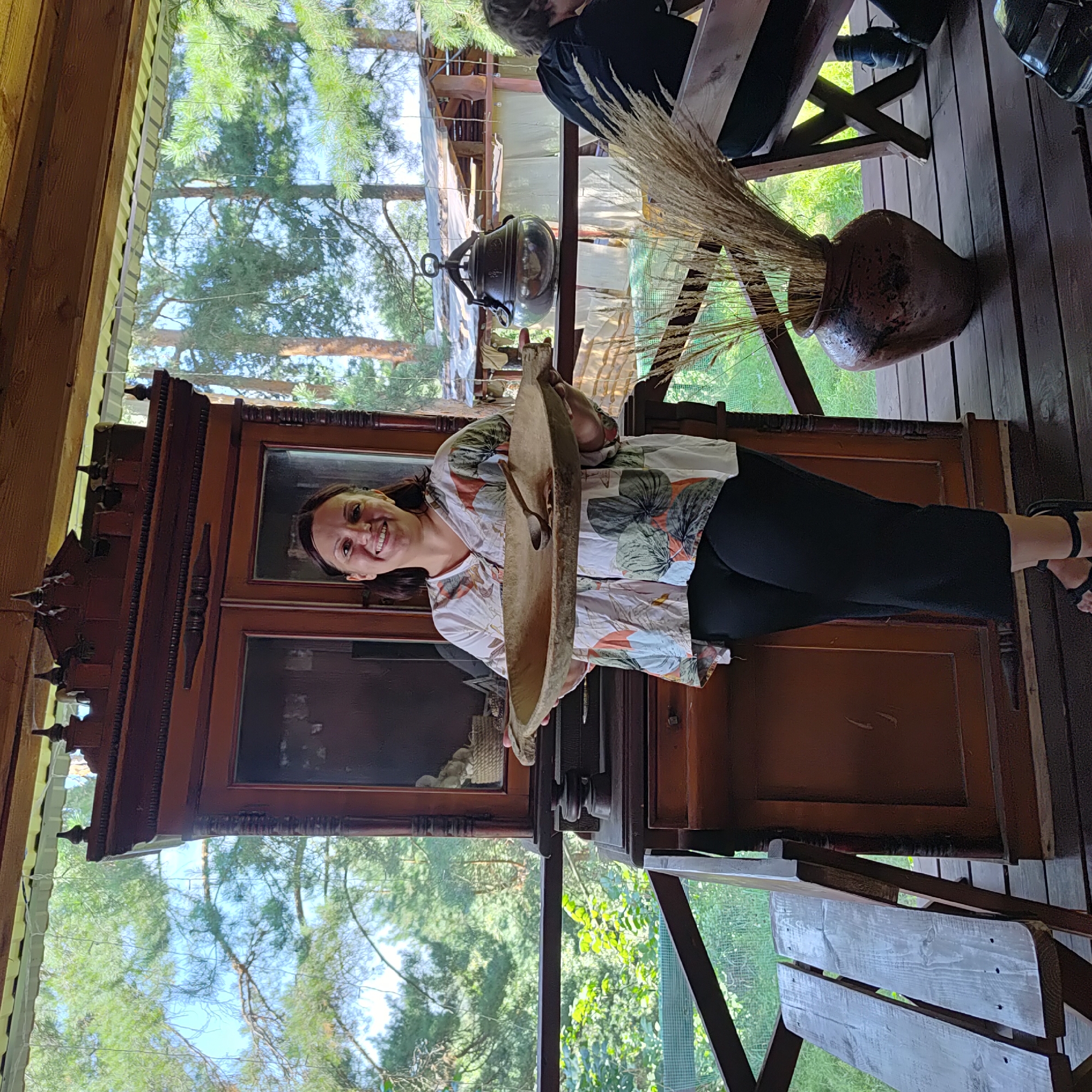 A woman with dark hair and a floral-patterned blouse and black pants stands on a wooden deck, holding a large wooden trough with a spoon inside. She is smiling at the camera. Behind her is a tall, dark wood cabinet with glass doors. The deck overlooks a wooded area with tall pine trees. To the right, a rustic shelter with a corrugated roof is visible, and a person in a black shirt is seated. A large ceramic vase filled with dried grass is on the deck to the right of the woman.
