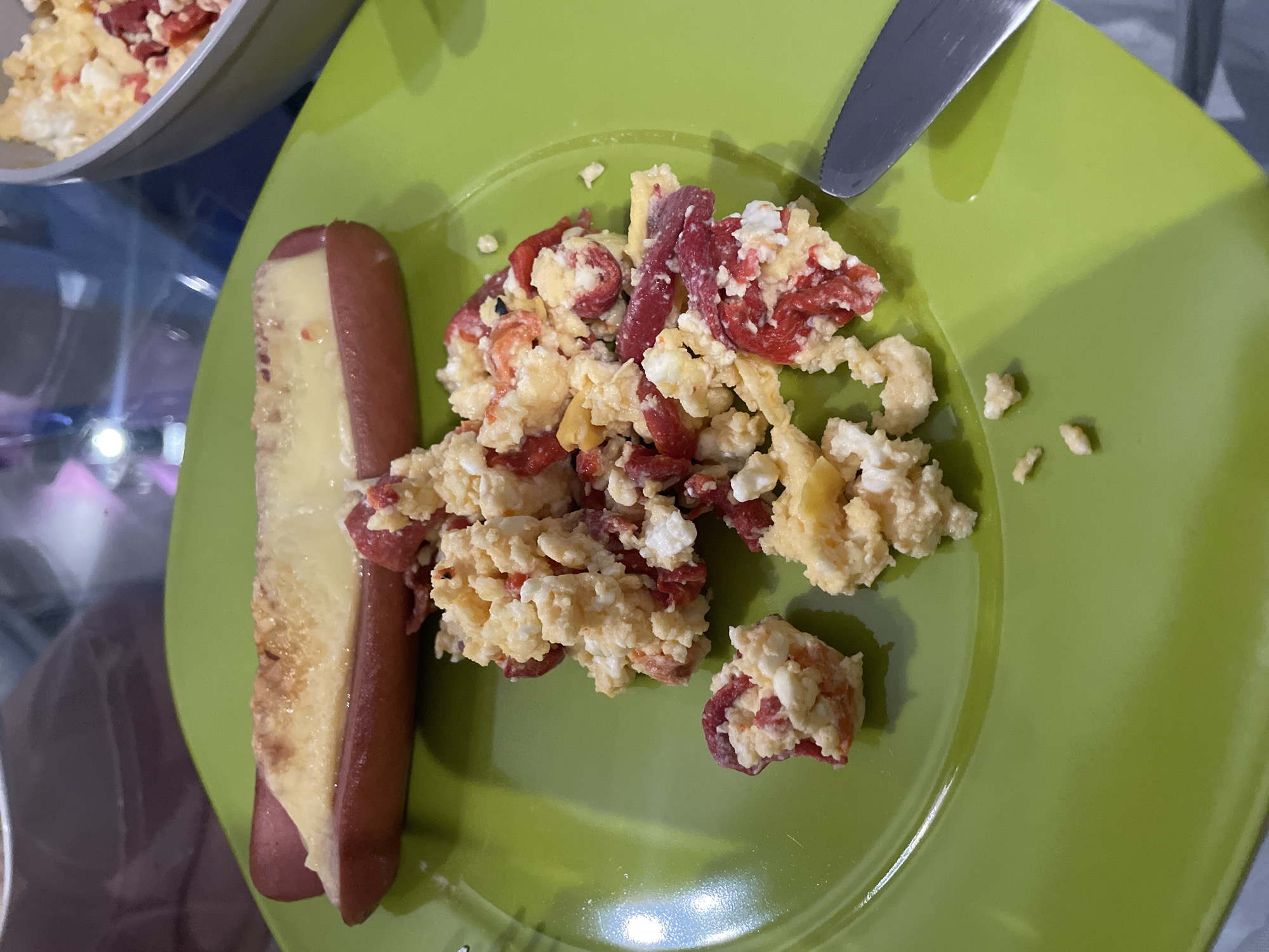 The image displays a close-up, overhead view of a meal served on a vibrant lime green plate. The main subjects are food items: a serving of scrambled eggs mixed with reddish ingredients, and a hot dog or sausage topped with a melted, light yellow substance, possibly cheese or an egg coating. A silver knife rests on the upper right edge of the plate.
In the upper left background, part of a grey bowl is visible, containing a similar mixture of scrambled eggs with red inclusions, suggesting additional portions or a different serving dish. The table beneath the plate appears to be made of glass, indicated by the blurred reflections and shapes visible through it. The overall setting is indoors, likely a dining area or kitchen, and the meal, consisting of eggs and sausage, suggests it is breakfast or brunch. The food appears freshly prepared, with the scrambled eggs having a chunky texture and the sausage or hot dog looking browned and topped. No specific location cues for Sofia, Bulgaria, are visible in the image itself, nor is there any visible text.