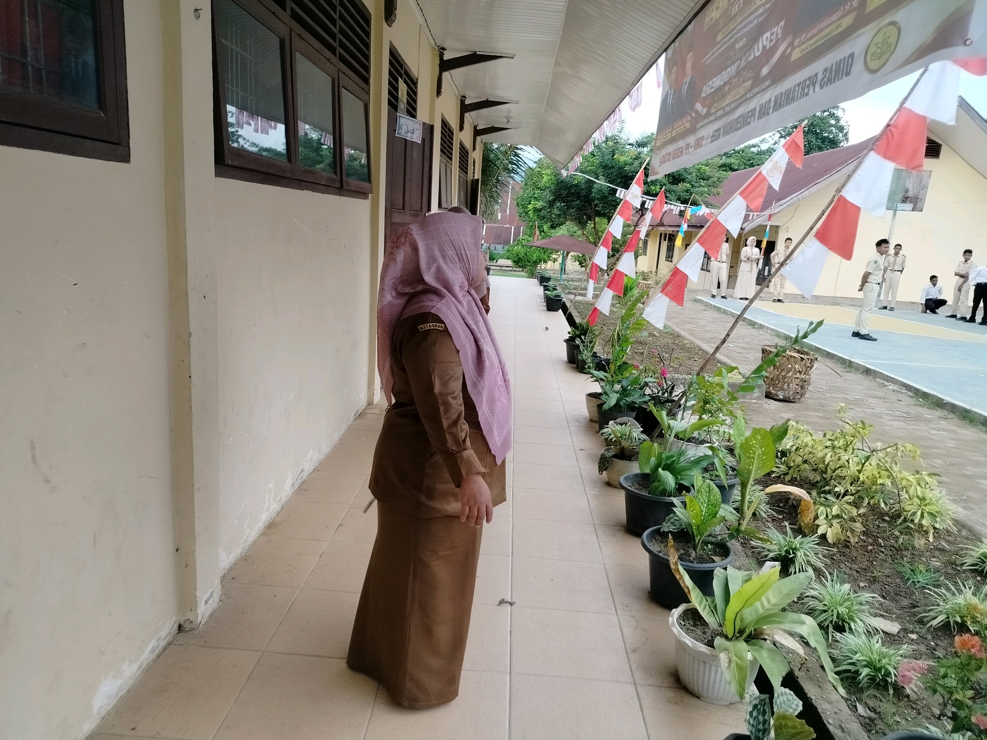 The image depicts an outdoor scene in what appears to be a school or public institution in Purwodadi, Indonesia. The foreground features a person wearing a brown uniform and a pink headscarf, standing on a tiled walkway. To the left, a beige-colored building with dark-framed windows is visible. Along the walkway, numerous potted plants and a flowerbed create a green border. In the background, several individuals in uniforms, possibly students, are gathered on a court area. Red and white Indonesian flags are displayed along poles, some attached to the building's awning, suggesting a national celebration or event. A large banner with text and images is hung from the awning. The overall environment suggests an institutional setting on a clear day, with elements of local culture and national pride evident through the flags and uniforms.