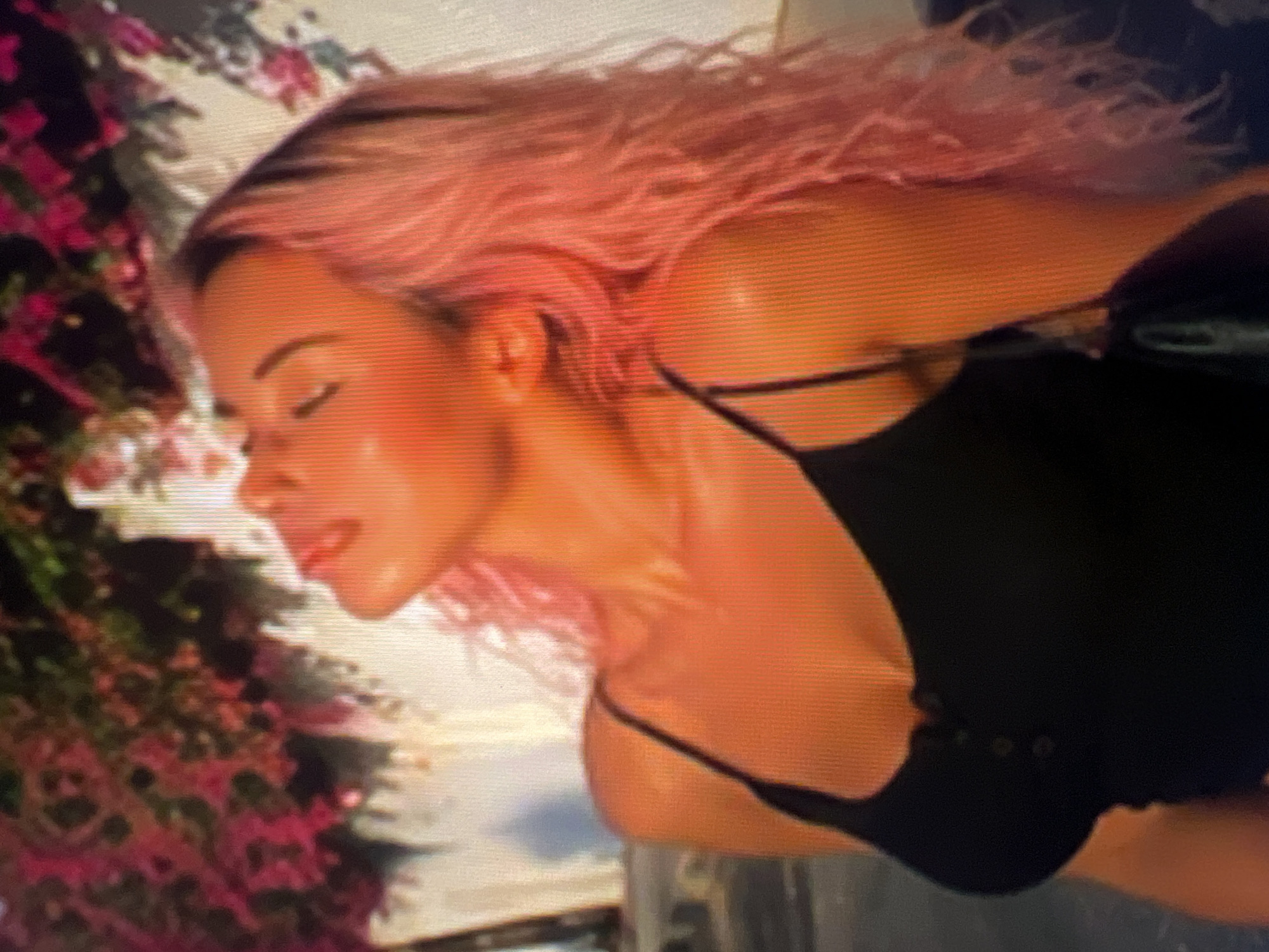 In an outdoor, sun-drenched environment with vibrant pink flowers in the background and a white, textured wall, a woman with bright pink, wavy hair is captured from the chest up. She wears a black, spaghetti-strap tank top with three buttons on the front. Her head is tilted back, eyes closed, and her face is illuminated by warm, golden light, suggesting a moment of relaxation or enjoyment. Her skin appears to have a dewy sheen. The background elements, including the foliage and wall, are slightly out of focus, drawing attention to the woman as the central subject.