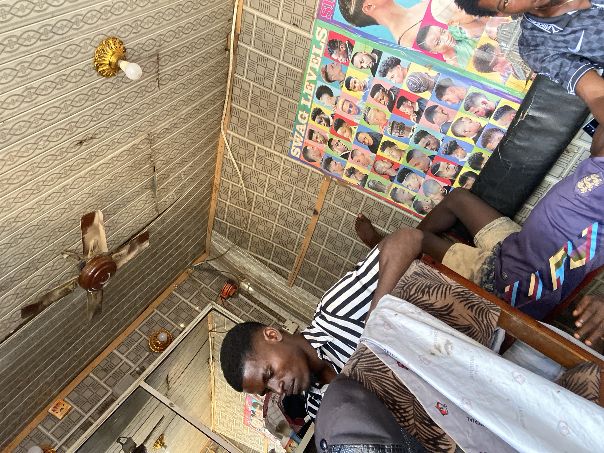 This media file depicts an indoor scene in Kuson, Nigeria, likely a barbershop or a similar establishment. The setting features a textured ceiling and walls, with a ceiling fan visible. A poster titled "SWAG LEVELS" featuring various hairstyles and men's portraits is prominently displayed on the wall, suggesting the context of grooming or fashion.
The primary subjects are three individuals, all appearing to be young men. One man in the foreground, wearing a black and white striped shirt, is leaning forward, his eyes closed and head bowed, appearing relaxed or perhaps asleep. Another individual, wearing a purple shirt with emblems, is partially visible in the lower right corner, with their hands resting on their legs. A third person's legs and feet are visible in the upper right portion of the image, wearing patterned shorts and a dark, possibly athletic, shirt.
Objects in the scene include a decorative golden light fixture with a light bulb attached to the ceiling, and what appears to be a roll of paper or fabric with some text on it. The lighting suggests it could be daytime, though the interior setting makes it difficult to ascertain the exact time of day or weather conditions. The overall mood appears to be casual and relaxed, with the individuals seemingly at rest within the establishment.