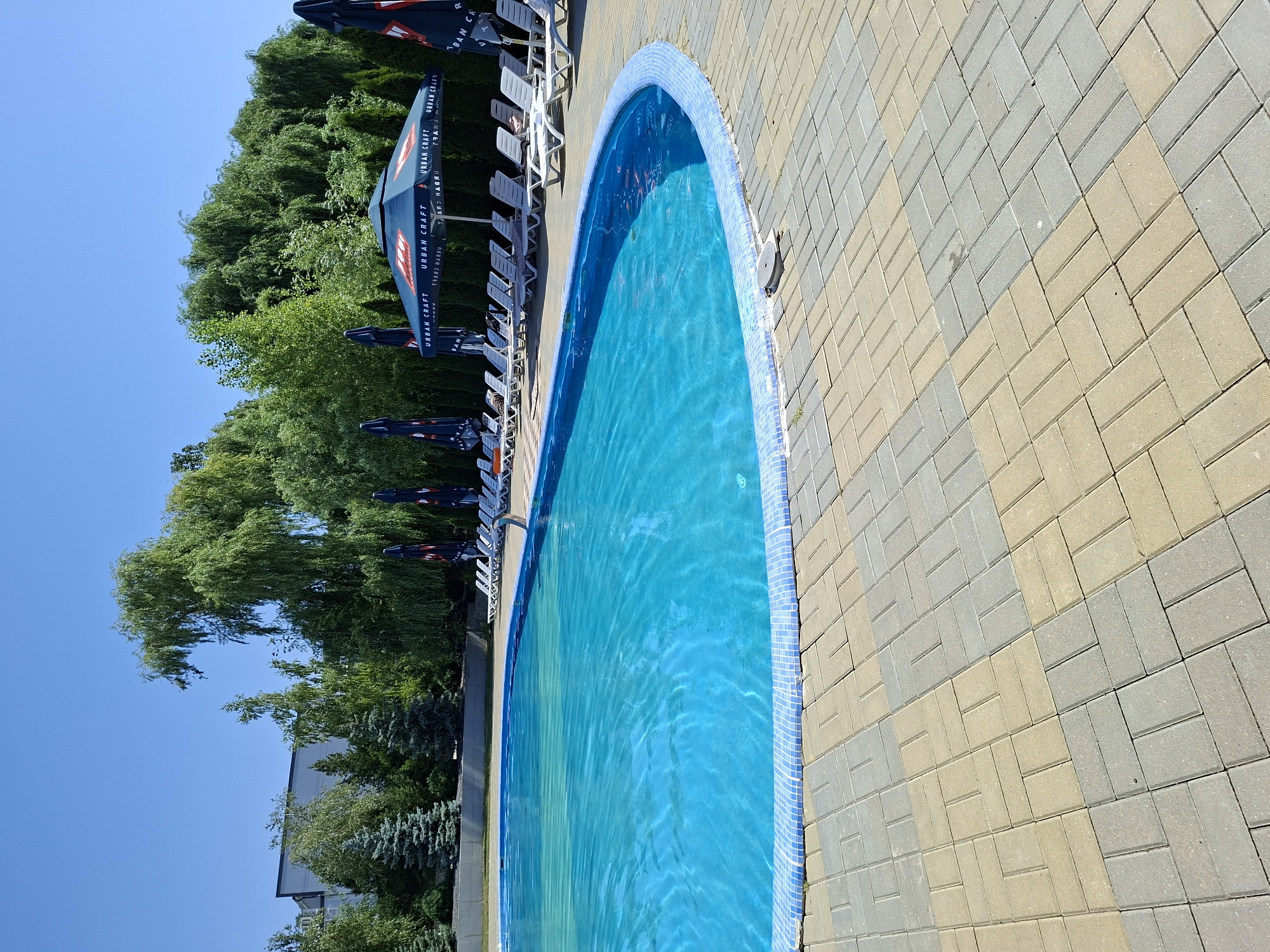 The image shows an outdoor swimming pool with bright blue water and a tiled edge. The pool area is paved with interlocking bricks in shades of tan and gray. Several white lounge chairs are lined up on the pavement, some with closed, dark blue umbrellas featuring a logo that appears to read "URBAN CRAFT". Lush green trees and some evergreen trees surround the pool, with a modern building visible in the background behind the trees. The sky is clear and blue. The location is identified as Costești, Moldova.
