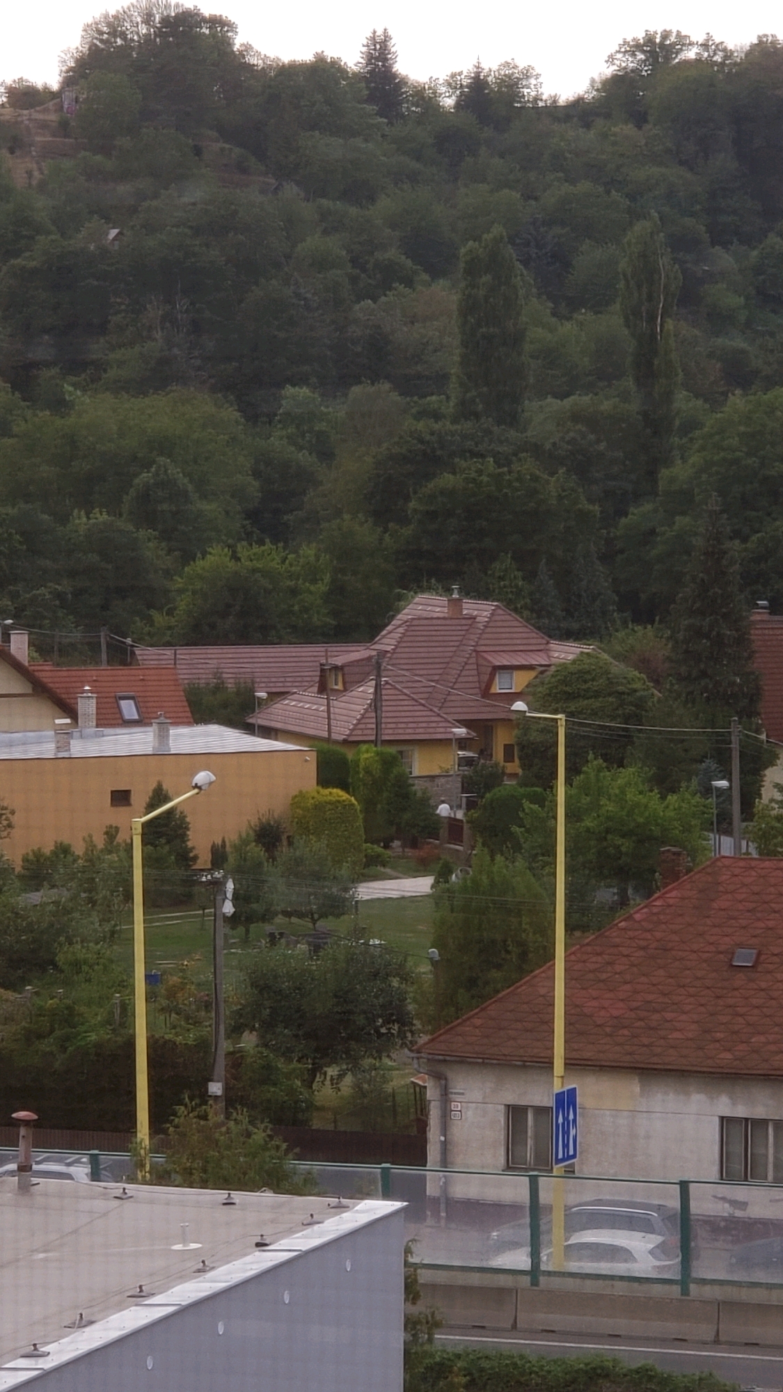 The image depicts a residential area in Prešov, Slovakia, with houses nestled amongst lush green trees. The foreground shows a portion of a modern building's grey flat roof. In the mid-ground, several houses with traditional red tile roofs are visible. One house, painted yellow, features stone accents around its base. A bright yellow street lamp stands prominently in front of it. Further back, a dense forest covers a hillside. The sky above is overcast. A white car is parked behind a barrier in front of one of the houses.