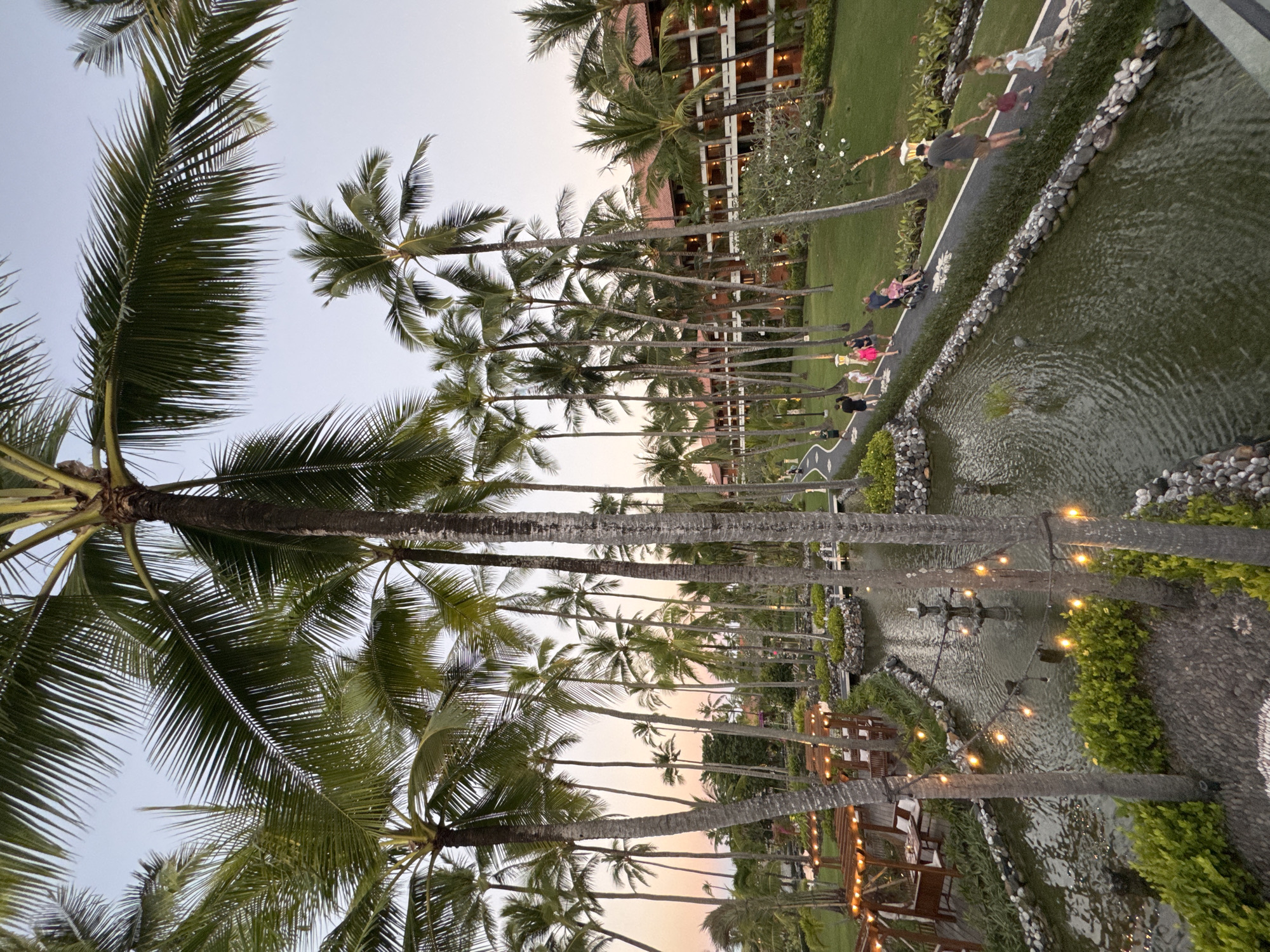 The image depicts a scene at sunset with palm trees lining a waterway in Kuta Selatan, Indonesia. A resort building with multiple lit windows is visible in the background. Along the waterway, people are walking on a pathway with decorative flower-shaped designs. A stream runs parallel to the path, and it is adorned with string lights. A fountain is in the water. The overall environment suggests a resort or leisure area within Kuta Selatan.