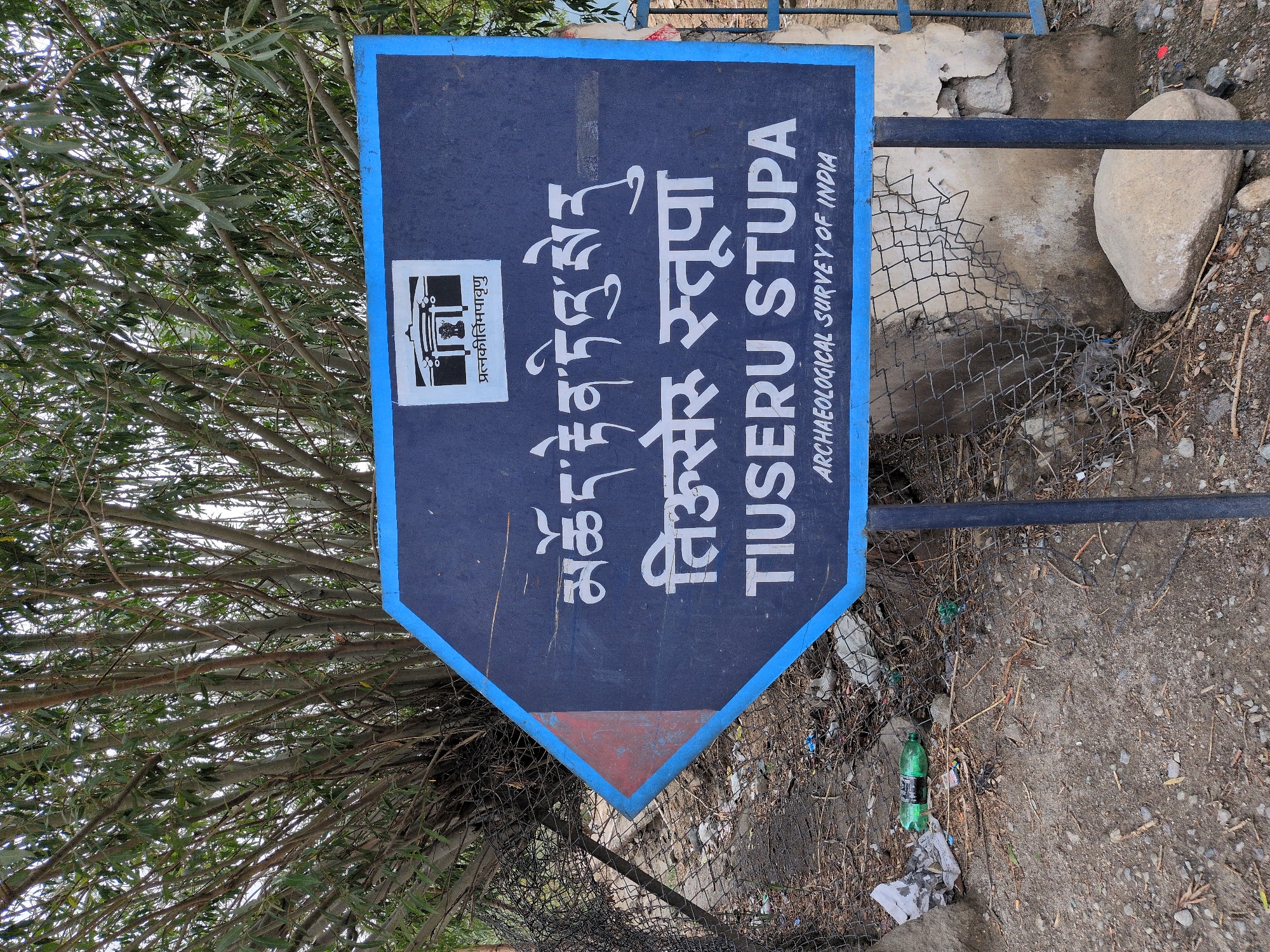 The image shows a directional sign in the city of Leh, India, pointing towards "Tiusseru Stupa". The sign is dark blue with white lettering in English and Tibetan script. Above the Tibetan script, a small building icon is depicted with text in Hindi below it. The sign is mounted on a pole. In the background, there is dense foliage of trees and a chain-link fence. The ground in front of the sign is littered with debris, including plastic bottles and paper. The sign is also weathered and has some scratches. The overall scene suggests a remote or historical site.