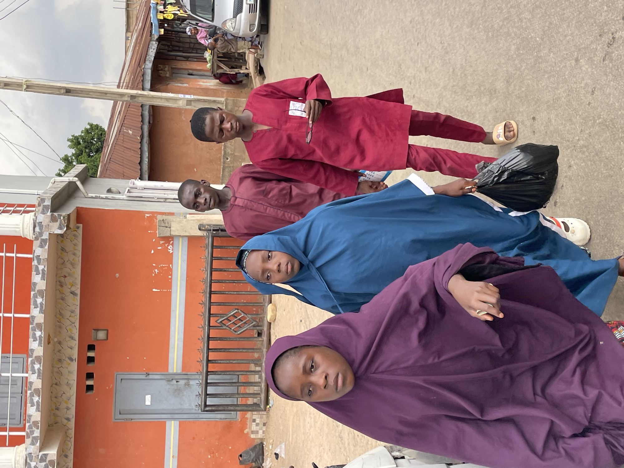 Four individuals are standing on a paved street in Jos, Nigeria, outside a building with an orange exterior. The building has a black metal gate and gray doors. The people are standing in a row, seemingly posing for a photo. The individual closest to the viewer is wearing a purple hijab. The second person is draped in a blue garment. The third and fourth persons are wearing maroon clothing. The fourth person is holding a pair of spectacles. A black plastic bag lies near the group. In the background, there are other buildings, including a vehicle and what appears to be a street vendor.