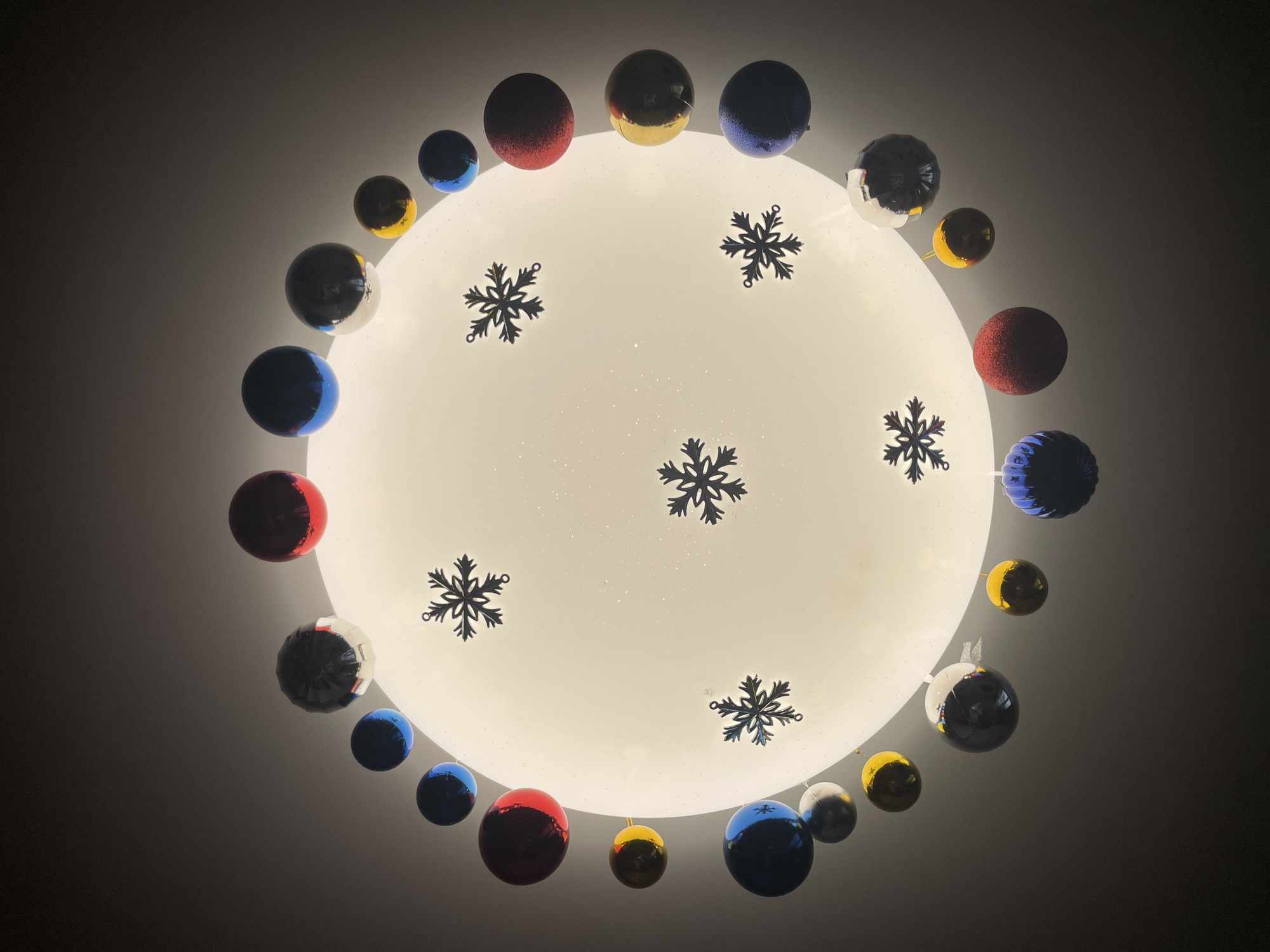 The image is a close-up, upward view of a ceiling light fixture adorned with Christmas ornaments. The light fixture itself is round and white, decorated with black snowflake designs. Around the perimeter of the light fixture are various colorful Christmas ornaments, including red, blue, gold, and silver balls. The scene is dimly lit, and the background is dark. There is no information in the image that indicates this is located in the city of Izhevsk, Russia.
