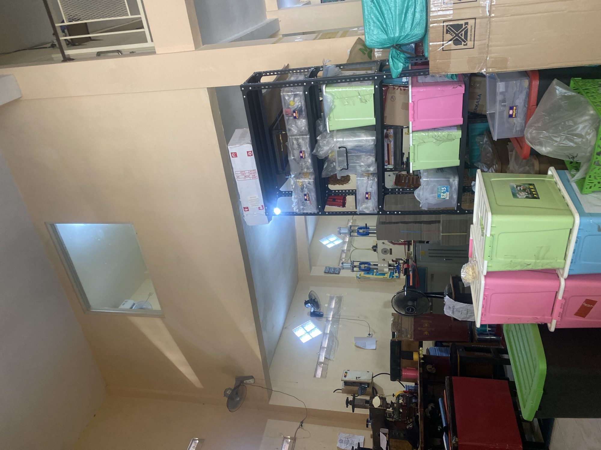 The image depicts an interior scene of what appears to be a workshop or storage area, likely located in Karangbong, Indonesia. The space is filled with shelves, storage containers, and machinery.
On the left side of the image, a red cabinet is visible, along with a desk and what looks like a piece of industrial equipment, possibly a stamping machine, with various tools and components on its surface. A fan is mounted on the wall above this area. Another fan is positioned on a desk in the middle of the room.
A large, black metal shelving unit dominates the right side of the image. It is stocked with numerous clear plastic bins and several brightly colored storage containers, including green and pink bins. Some bins contain what appear to be various small items, possibly parts or supplies. A large, green tarp is also visible to the right of the shelving unit.
In the background, more machinery and equipment can be seen, including what appears to be a large gray industrial machine. Two windows with diamond-patterned glass are visible on the wall, letting in natural light. A small window is also present higher up on the wall, offering a view of what seems to be an upper level or another room. The walls are painted a light beige or cream color, and the ceiling is also light-colored. The overall impression is of a functional, cluttered workspace.