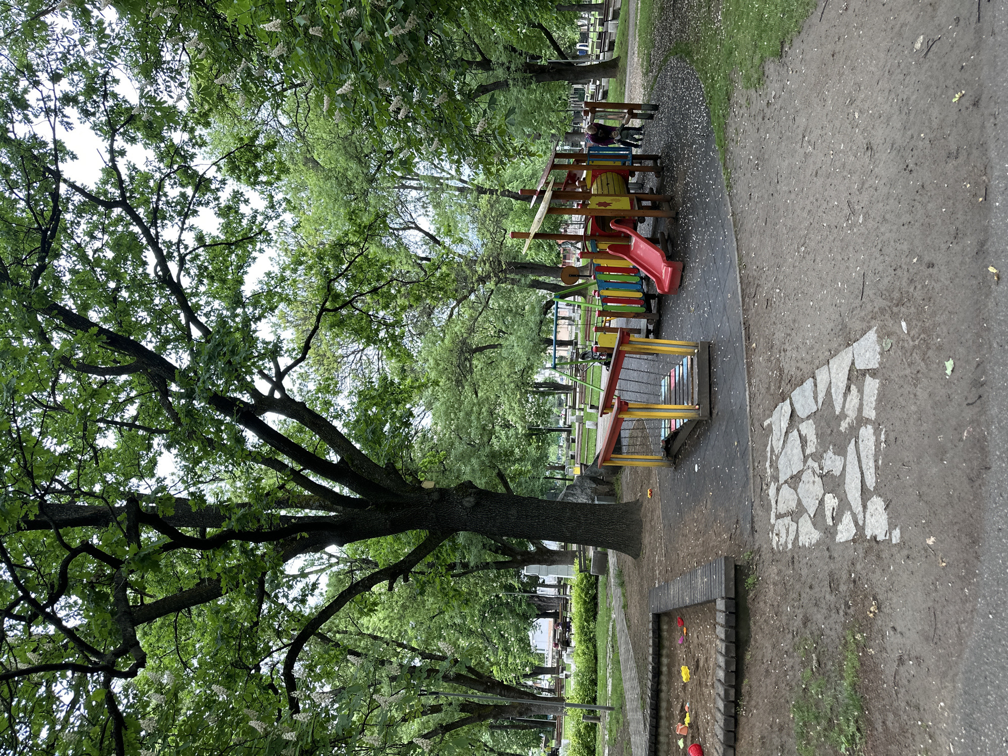 The image shows a playground in Montana, Bulgaria. A large tree with sprawling branches occupies the left side of the frame, its leaves creating a canopy overhead. To the right is a children's play structure with a red slide, colorful bars, and wooden platforms. In the background, more trees line the pathway. The ground is a mix of paved areas, dirt, and patches of grass. A stone mosaic pattern is visible in the foreground. There's also a sandbox area bordered by wooden planks, containing colorful plastic toys. People are seated on a bench near the play structure.