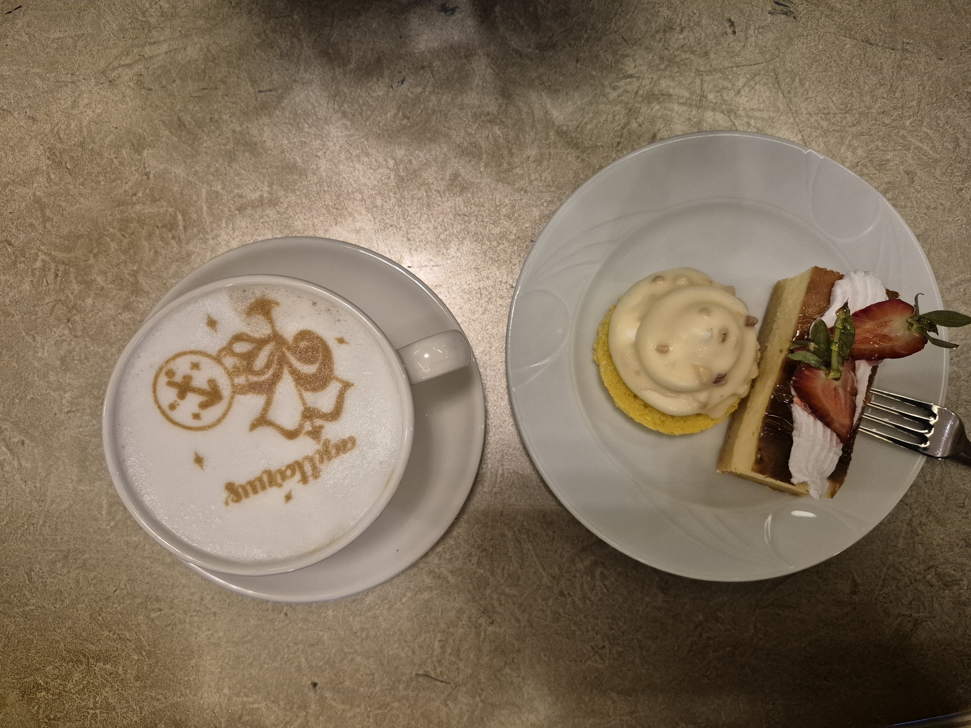 The image shows a cappuccino on a saucer next to a plate with two desserts, likely photographed indoors on a table with a textured surface. The cappuccino has a "Sagittarius" symbol and the name "Sagittarius" written in a decorative style on the foam. The plate contains a round dessert with a yellow base and a light-colored topping, and a slice of cake decorated with strawberries and whipped cream, with a fork resting on the plate. The specific location of the photo in Alanya, Turkey cannot be ascertained from the image.