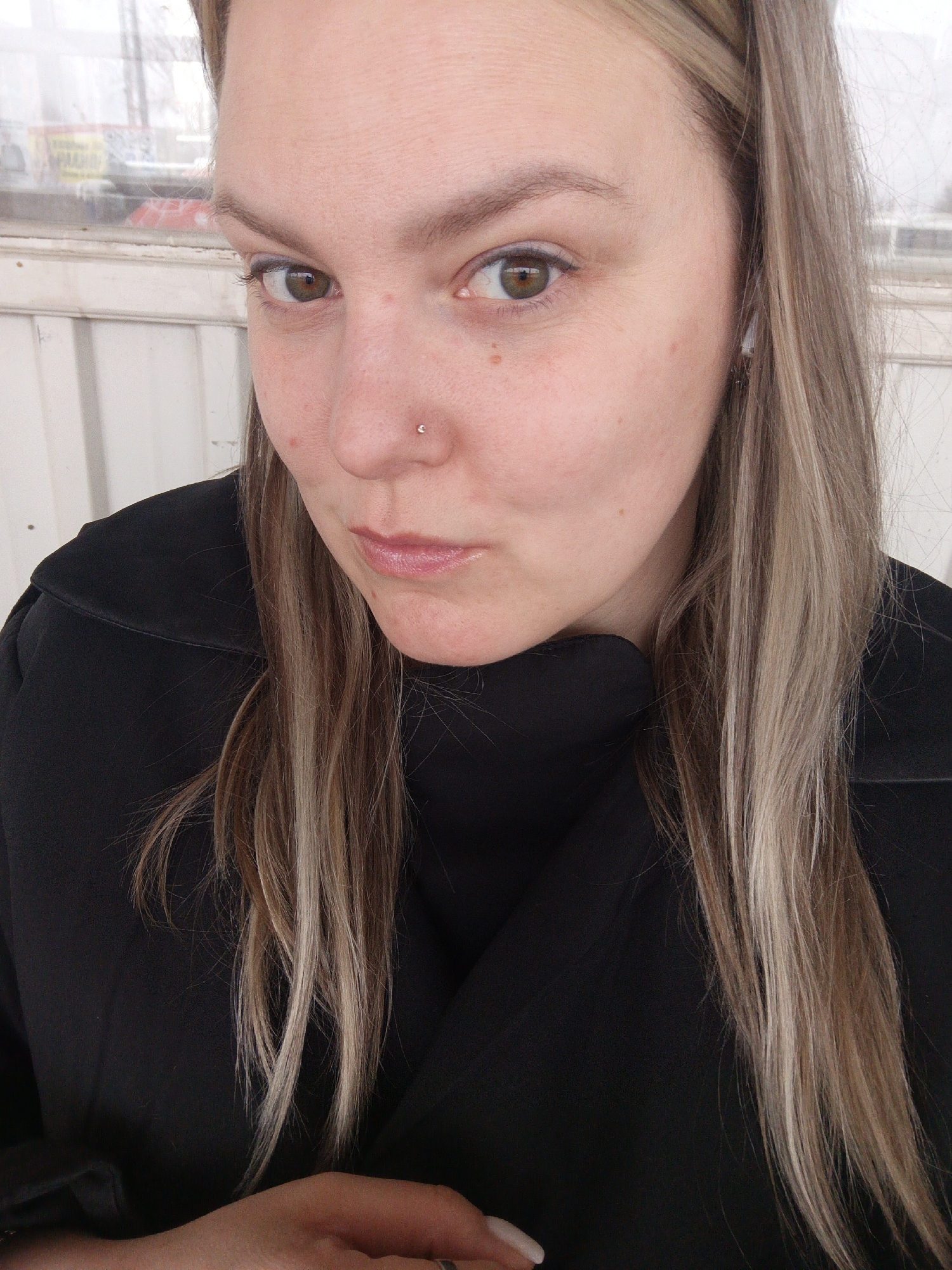 A woman with light brown and blonde highlighted hair is facing the camera. She has a nose piercing on her left nostril. She's wearing a black top. Her facial expression is a slight pursing of the lips. The background appears to be outdoors, possibly near a building with window frames and blurry signs. The location is indicated as Chaikovsky, Russia.