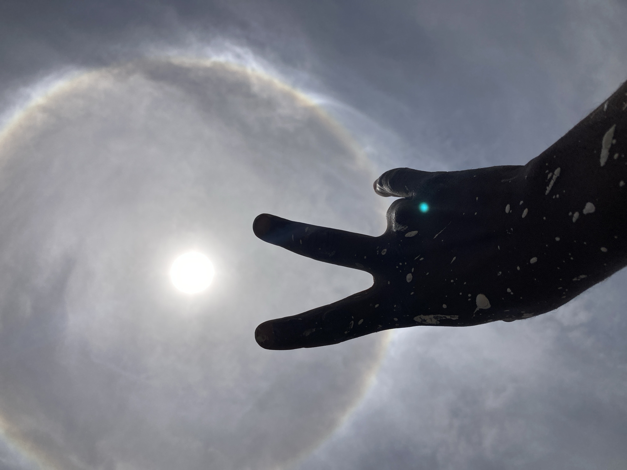 The image presents an upward view of a bright, cloudy sky featuring a striking solar halo. The sun, a brilliant white orb, is prominently positioned towards the left-center, surrounded by a large, circular, and faintly iridescent halo, indicative of ice crystals in the atmosphere.
Extending from the right side of the frame is a human hand, silhouetted against the intense light. The hand appears dark, possibly due to backlighting or a dark glove, and is covered with scattered white specks, resembling paint or dust. The hand's gesture shows the index, middle fingers, and thumb extended, while the ring and pinky fingers are curled. A small, greenish-blue lens flare is visible near the tip of the middle finger, an artifact of the camera's interaction with the direct sunlight.
The overall scene captures a meteorological phenomenon during daytime, likely mid-day given the sun's high position and intensity. The presence of the solar halo suggests specific atmospheric conditions with cirrus clouds. The photograph, taken in Jos, Nigeria, focuses on the interaction between a human element and a natural spectacle, emphasizing the wonder of the solar halo. There is no visible text in the image.