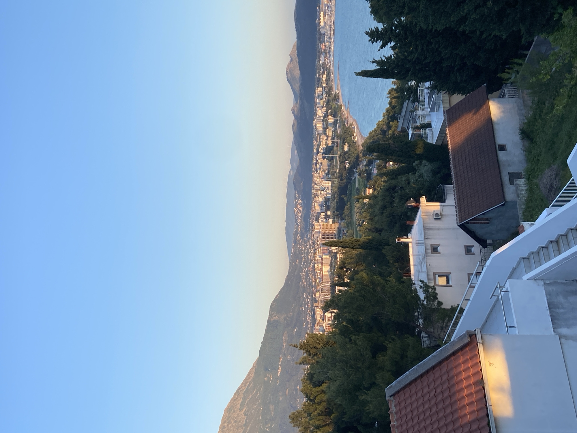This image depicts a coastal cityscape in Šušanj, Montenegro, likely taken from an elevated position. The scene includes a clear, light blue sky dominating the left side of the frame, transitioning to a lighter hue towards the center horizon. Below the sky, a mountainous landscape runs horizontally, with a cluster of buildings scattered at its base. These buildings form the urban area of Šušanj, extending along the coastline. The Adriatic Sea appears as a blue expanse bordering the city. A mix of evergreen trees and other vegetation dots the terrain between the city and the viewpoint. In the foreground, architectural elements like roof tiles, white walls, and staircases suggest a building or terrace from which the photo was taken. No people are visible in the frame. The lighting indicates either sunrise or sunset, casting a warm glow on the buildings and landscape.