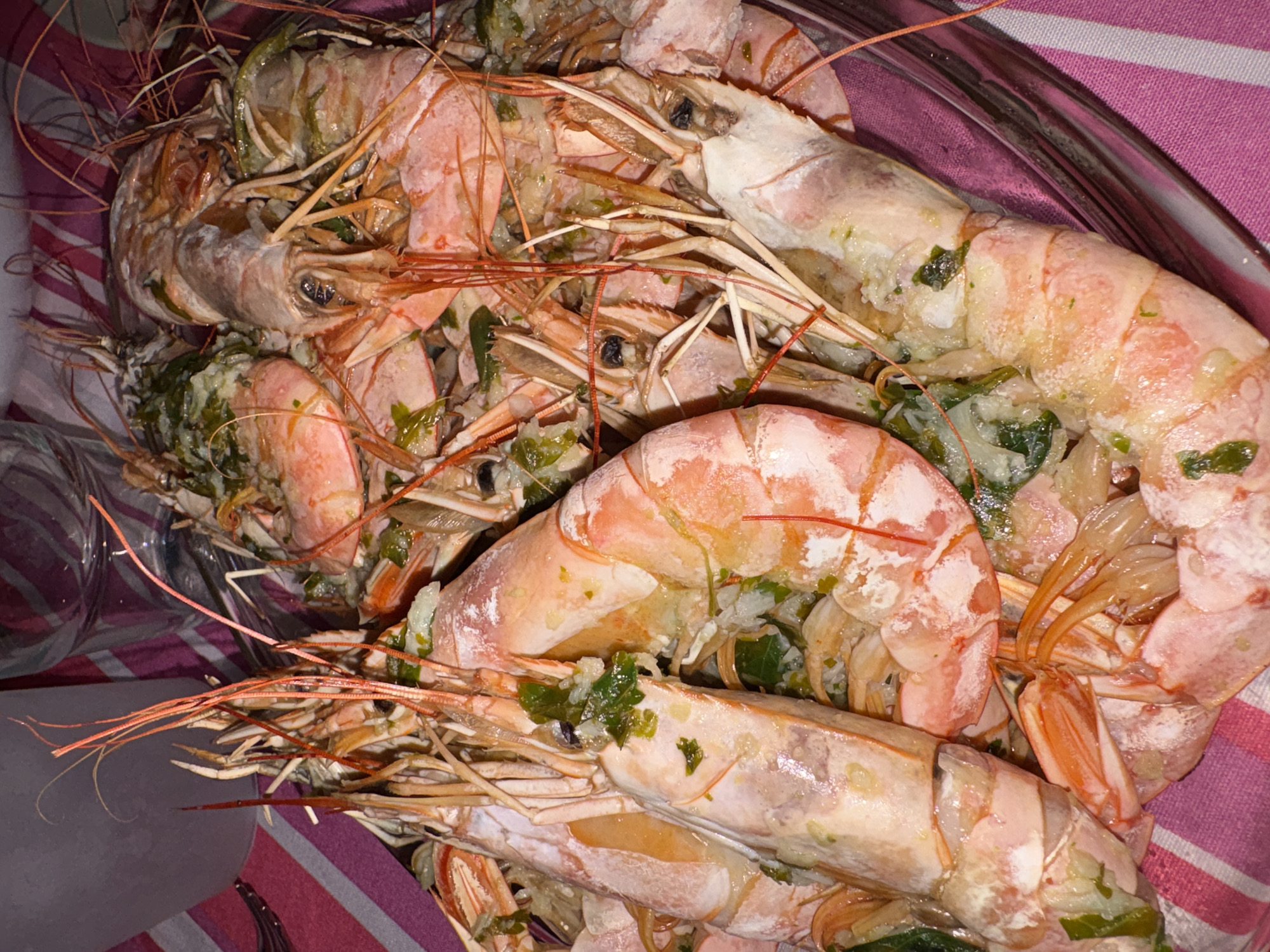 A close-up, high-angle shot reveals a generous serving of cooked prawns or large scampi piled in a clear glass bowl. The crustaceans are reddish-pink, indicative of being cooked, with their shells and long antennae mostly intact. They are visibly seasoned with finely chopped green herbs, likely parsley or cilantro, and possibly garlic or onion, embedded within and atop their shells, suggesting a flavorful preparation. Some liquid or oil is visible, indicating they might be marinated or served with a sauce.
The glass bowl rests on a surface covered with a pink and white striped fabric, possibly a tablecloth or napkin, which serves as a backdrop. Part of a clear drinking glass is visible in the lower left portion of the frame, hinting at a dining setting. No people are visible, and no specific activity beyond the presentation of the food is depicted. The lighting appears to be indoor, and neither the time of day nor the weather can be determined from the image. There is no visible text. The setting for this meal is in Urbanización Encinares De Alcolea, Spain.