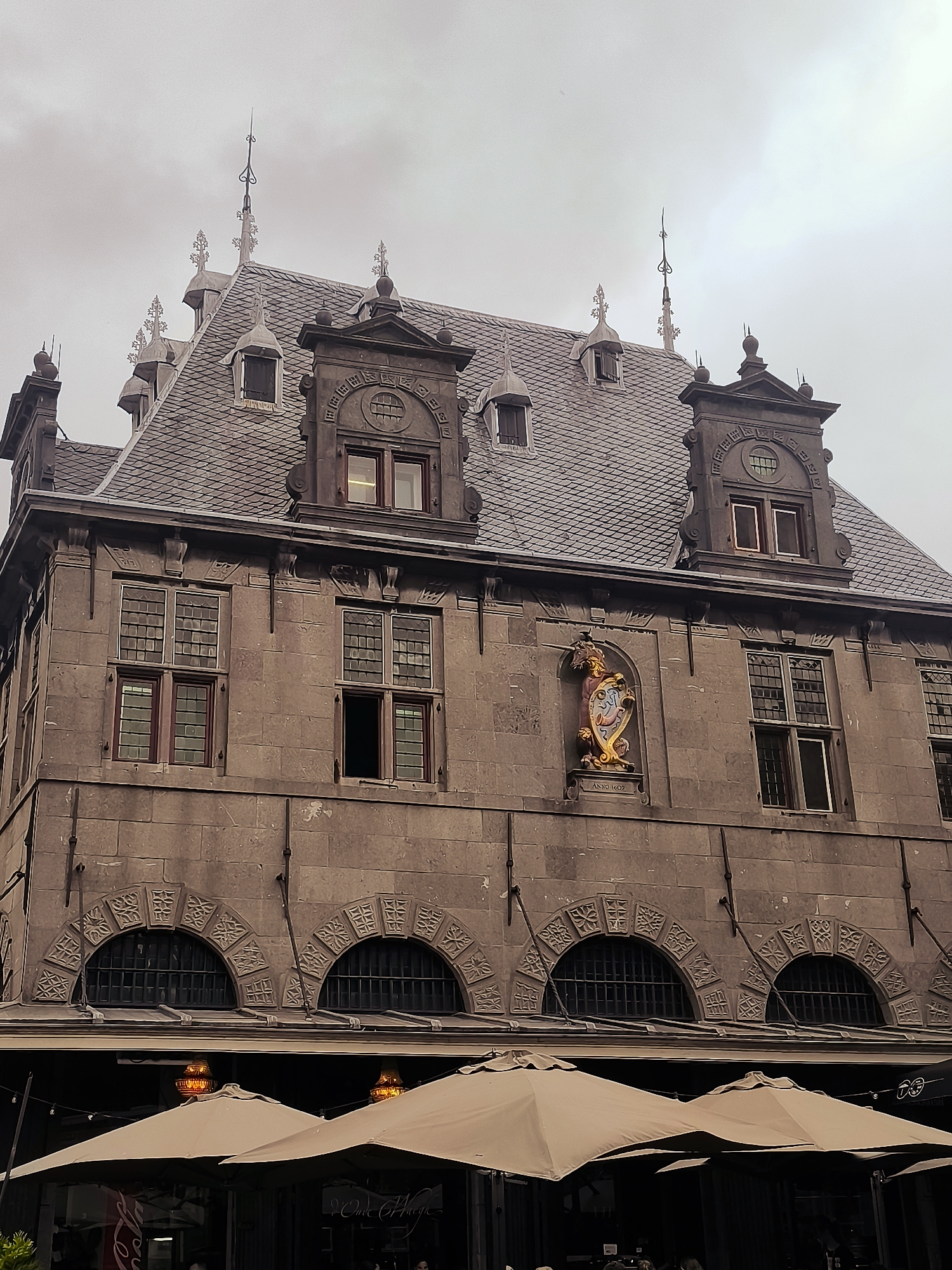 This is a medium-shot, eye-level, exterior photograph of the Westfries Museum in Hoorn, Netherlands, taken on an overcast day. The museum is a historic building with a grey, tiled, mansard roof featuring several dormer windows and decorative finials. The facade is constructed of stone blocks, with numerous windows that have multiple panes and dark wooden frames. Above the ground floor, which is partially obscured by outdoor seating with large, light-colored umbrellas, there are decorative arched windows with intricate stonework. A prominent feature on the facade is a colorful coat of arms mounted in a niche, with "ANNO 1602" inscribed below it. The overall tone of the image is muted due to the overcast sky and the monochromatic color palette of the building.