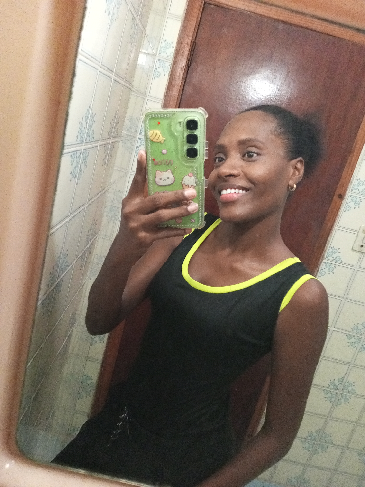 A young, dark-skinned woman is taking a selfie in a bathroom. She is holding a smartphone with a green case decorated with cartoon characters in her left hand. She is wearing a black tank top with yellow trim. Her hair is pulled back. She is smiling. The bathroom has patterned tiles on the walls and a brown wooden door. The location is Palo Negro, Venezuela.