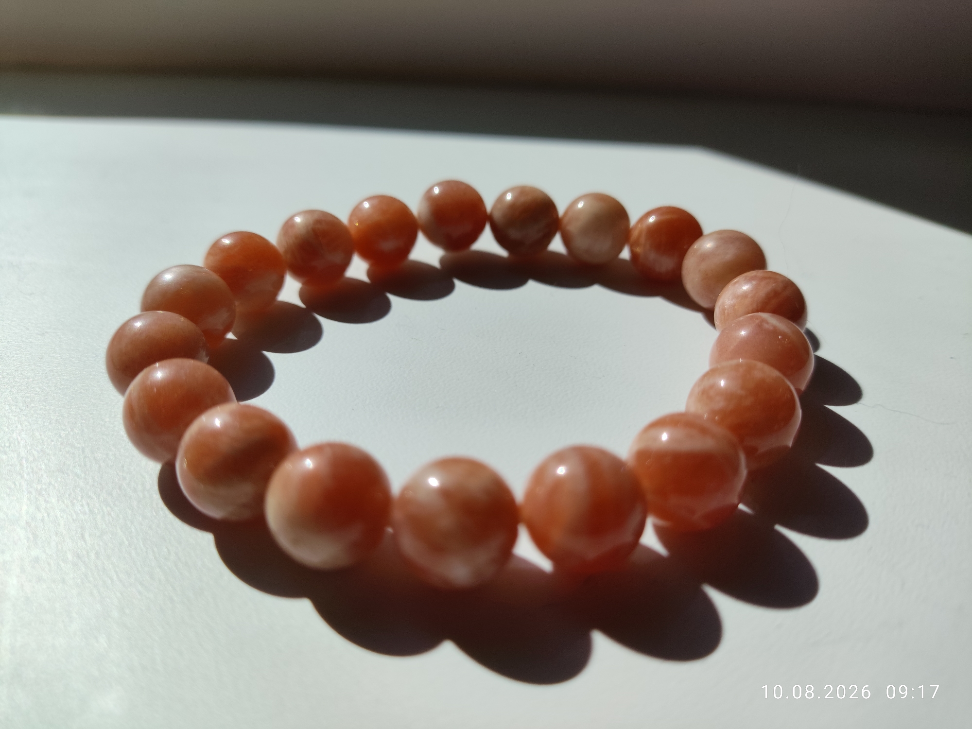 The image shows a bracelet made of round, orange-pink beads. The bracelet is laid out in a circular pattern on a white surface, casting shadows from the light source, which appears to be natural sunlight from the upper left. The beads have a smooth, polished finish and exhibit some variations in color and subtle internal patterns. The surface is clean and evenly lit, with a hint of texture. The overall scene is simple and uncluttered, focusing entirely on the bracelet. The image was taken on August 10, 2026, at 09:17. There is no indication of the city of Pavlodar, Kazakhstan, or any other specific location.