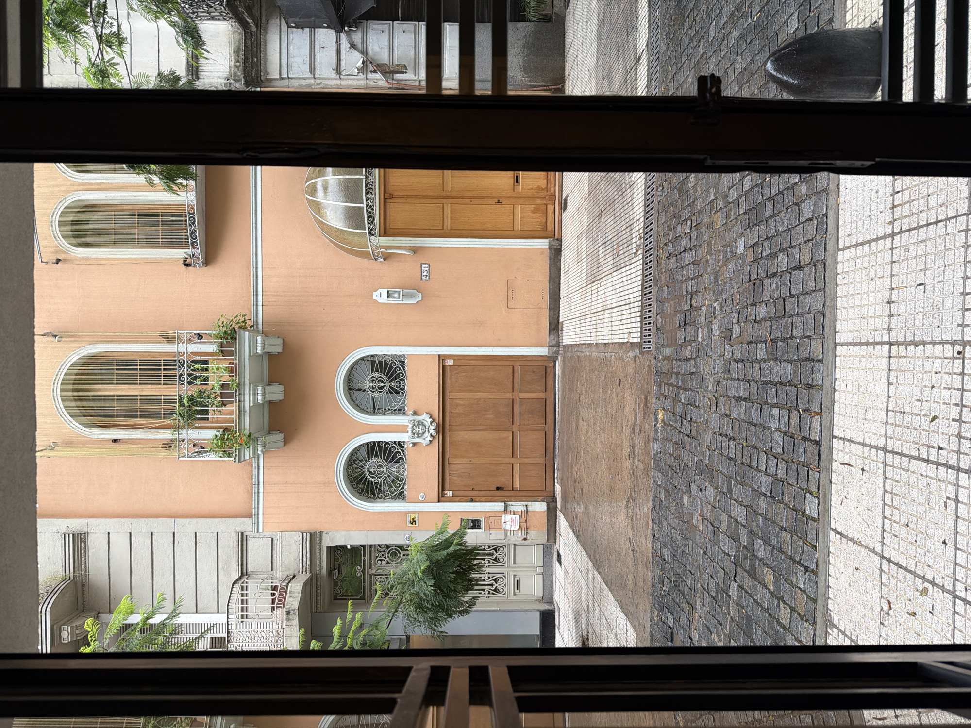 A vertical shot of an exterior view from indoors, potentially from a window, shows a street scene in Buenos Aires, Argentina. The predominant features include architecture with salmon-colored facades and a diverse pattern of pavement. Buildings directly across the street exhibit a mix of architectural styles, with some buildings having ornate window grills and others having modern door styles. There is a contrast in street paving materials, one area covered with interlocking gray stones, and another section with white tilework. The perspective suggests a view from an elevated position, looking down onto the street. The image is confined by what appear to be parts of the window frame and other interior elements at both top and bottom, creating a framed composition of the urban landscape.