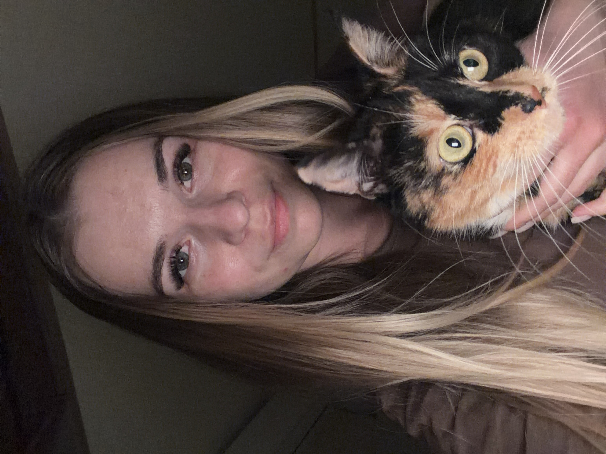 A woman with long blonde hair is holding a calico cat close to her face. The woman's face is partially visible, with visible makeup on her eyelashes. The cat has black, orange, and white fur, and its eyes are wide and yellow. The background is dark, suggesting an indoor location. The city of Unknown, Unknown is not discernible from the image.