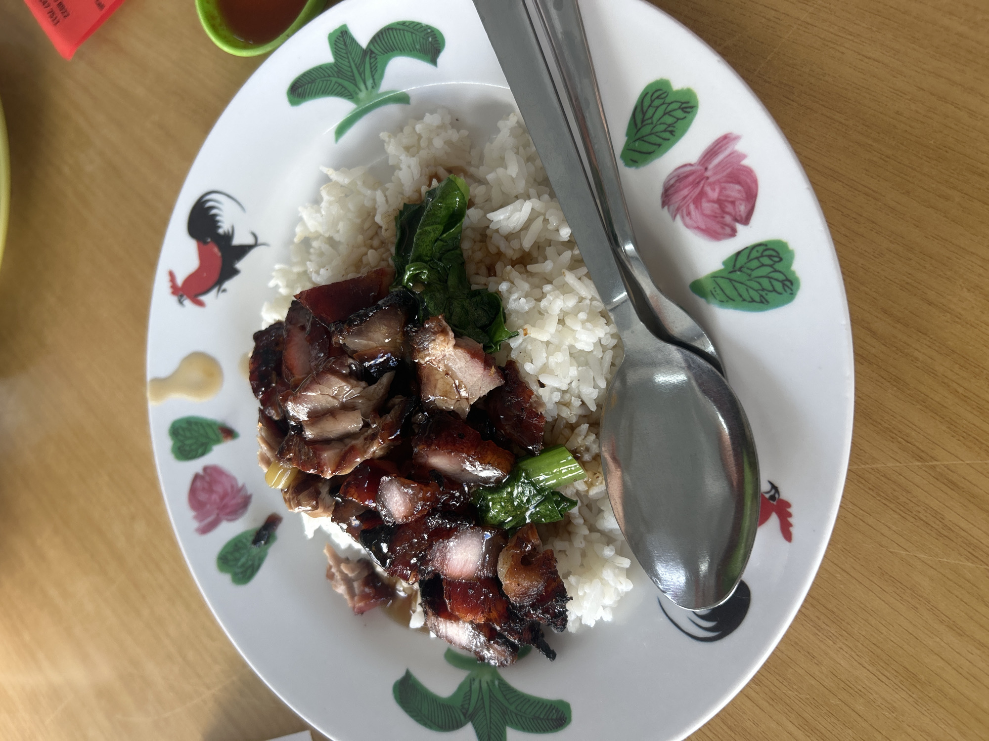 A close-up overhead shot shows a plate of roasted red pork with rice and greens, a common dish in George Town, Malaysia. The plate, adorned with rooster and floral motifs, rests on a wooden table. A spoon and fork are partially visible in the foreground, suggesting the meal is ready to be eaten. The lighting is natural, casting a warm glow on the food and table. The image focuses on the culinary experience, highlighting the texture and glistening appearance of the roasted pork.