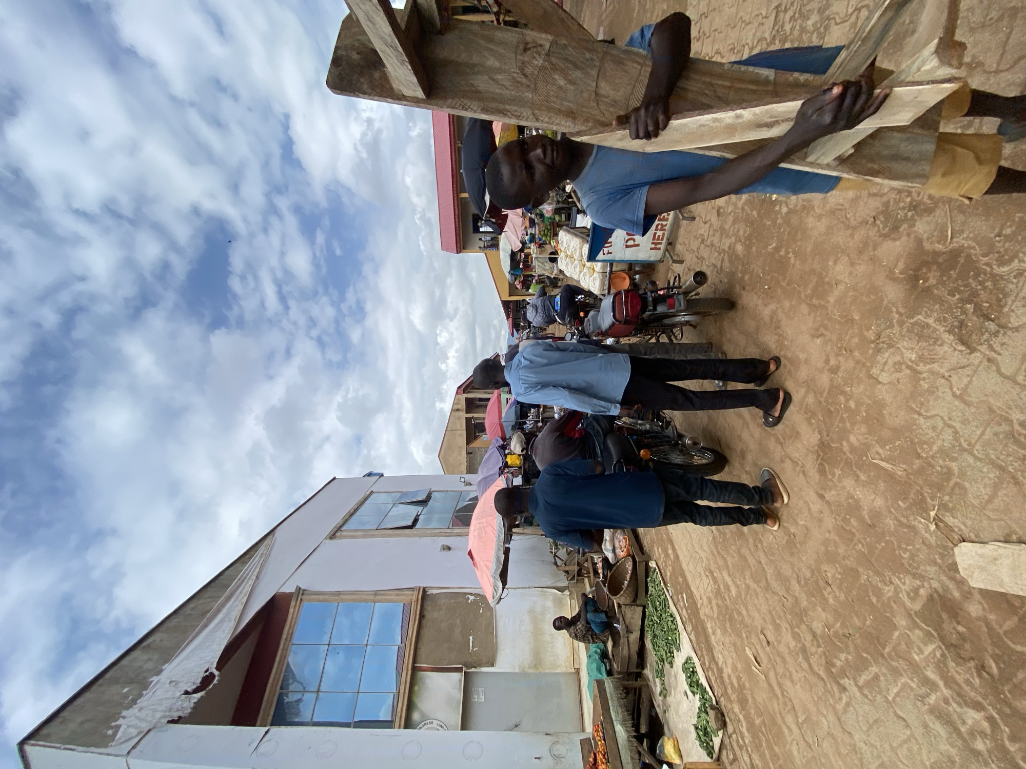 The image depicts a bustling outdoor market scene in Kafanchan B, Nigeria, under a cloudy sky. In the foreground, a young boy in a blue t-shirt and yellow shorts carries a large wooden frame. To his left, a man in a blue shirt and black trousers walks away from the camera, while another man in a dark blue shirt stands beside a motorcycle. Behind them, several other people are visible, some on motorcycles and others near market stalls. The market features various goods, including vegetables laid out on a mat, and numerous umbrellas providing shade. In the background, there are buildings, some appearing to be shops or market structures. The ground is a mixture of dirt and what looks like paving stones. The overall impression is one of a lively, active marketplace with a mix of commerce and pedestrian activity.