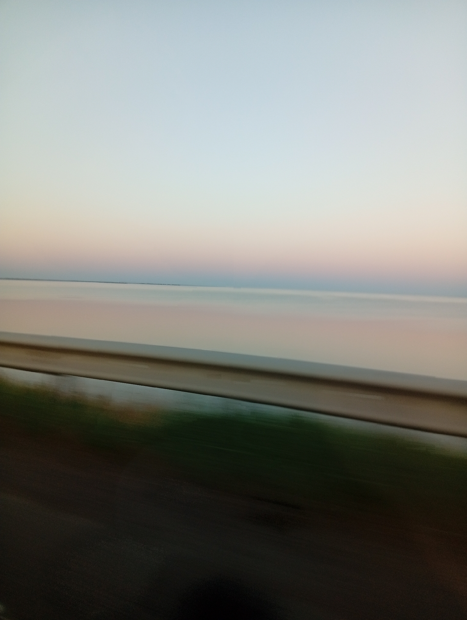 The image captures a serene landscape at what appears to be either sunrise or sunset, with a gradient of soft colors in the sky. A calm body of water stretches across the horizon, reflecting the sky's muted hues. In the foreground, a metallic guardrail runs horizontally across the frame, suggesting the vantage point is from a road or elevated pathway. To the left of the guardrail, a blurred band of green and dark brown indicates vegetation or the ground, also exhibiting motion blur from movement. The overall impression is one of peacefulness and a transition between day and night. The location is identified as Unknown, Unknown.