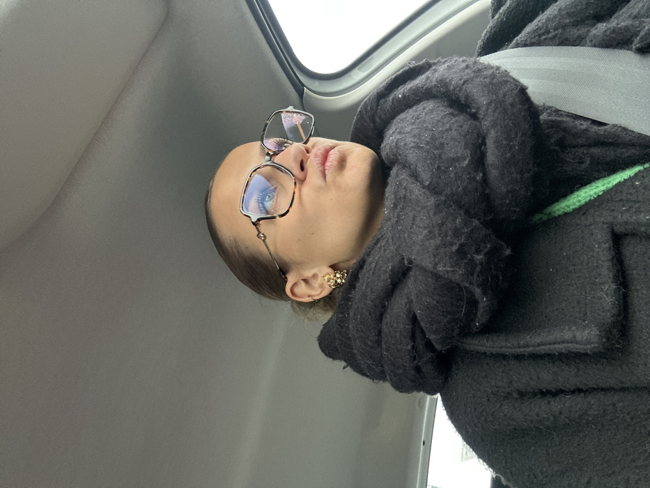 This image depicts a woman seated inside a vehicle in Saint-Cyprien, France. The woman, positioned from the chest up and angled slightly to her right, wears large, square-framed tortoiseshell eyeglasses, revealing her blue eyes. Her light brown hair is pulled back. She displays a gold-toned, ornate earring in her visible left earlobe, accompanied by a smaller stud piercing higher on the ear. Her lips are pursed forward. She is dressed in a thick, textured black coat and a voluminous black knitted scarf wrapped around her neck and lower face. A segment of a bright green knitted garment or strap is visible beneath her coat on her left side. A dark gray seatbelt is fastened across her chest. The vehicle's light gray headliner is visible above her, and a light-colored window frame is partially visible on the far right.