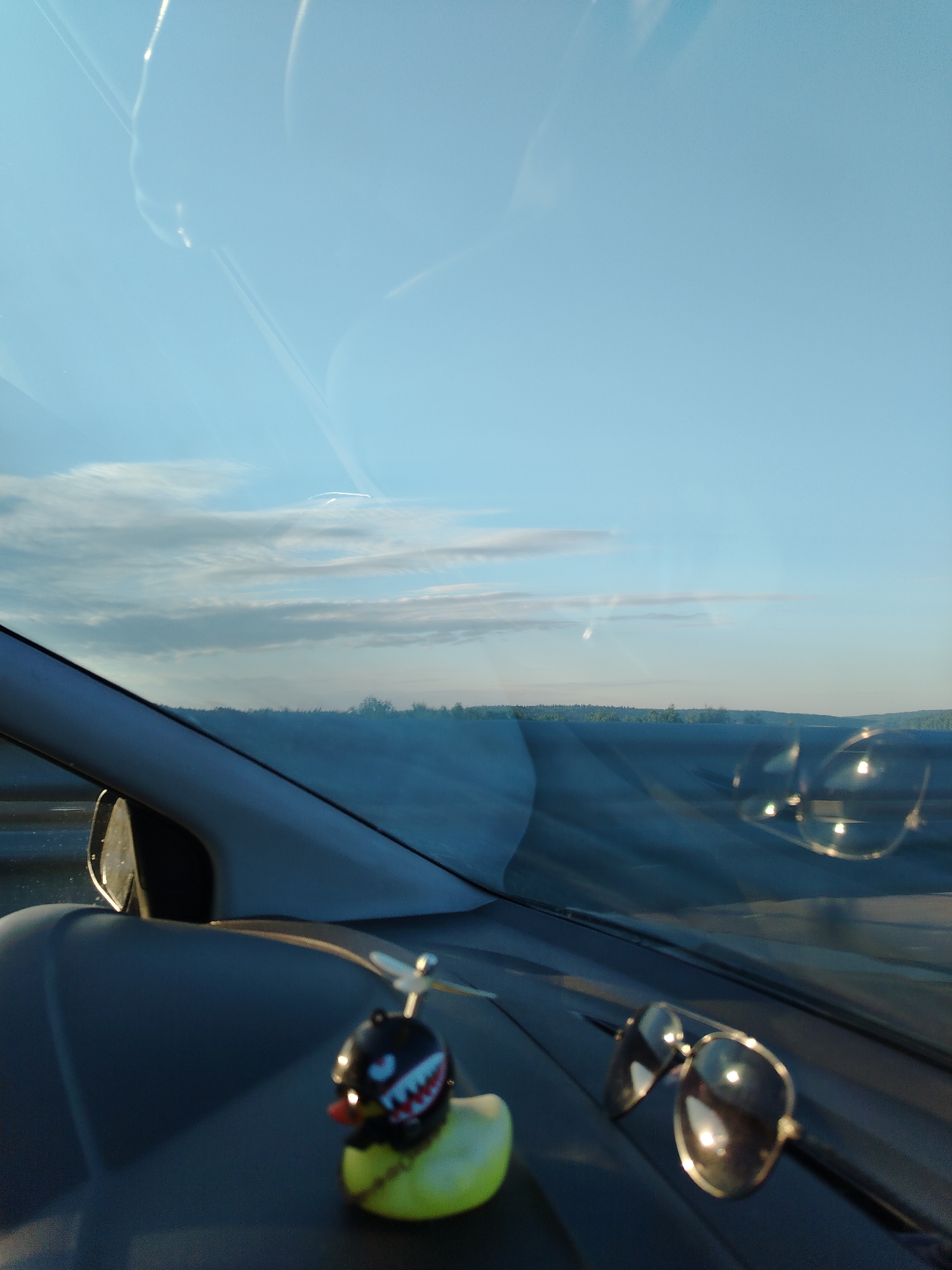 The image is taken from inside a vehicle, likely a car, looking out through the windshield. The sky is visible with light blue and some clouds. In the foreground, on the dashboard, there's a yellow rubber duck wearing a black helmet with a propeller on top and a pair of sunglasses are present. The location is not definitively identifiable as Bereznyaki, Russia, as there are no obvious landmarks or other visual cues that would indicate the specific geographic location. The landscape visible in the distance appears to be a line of trees.