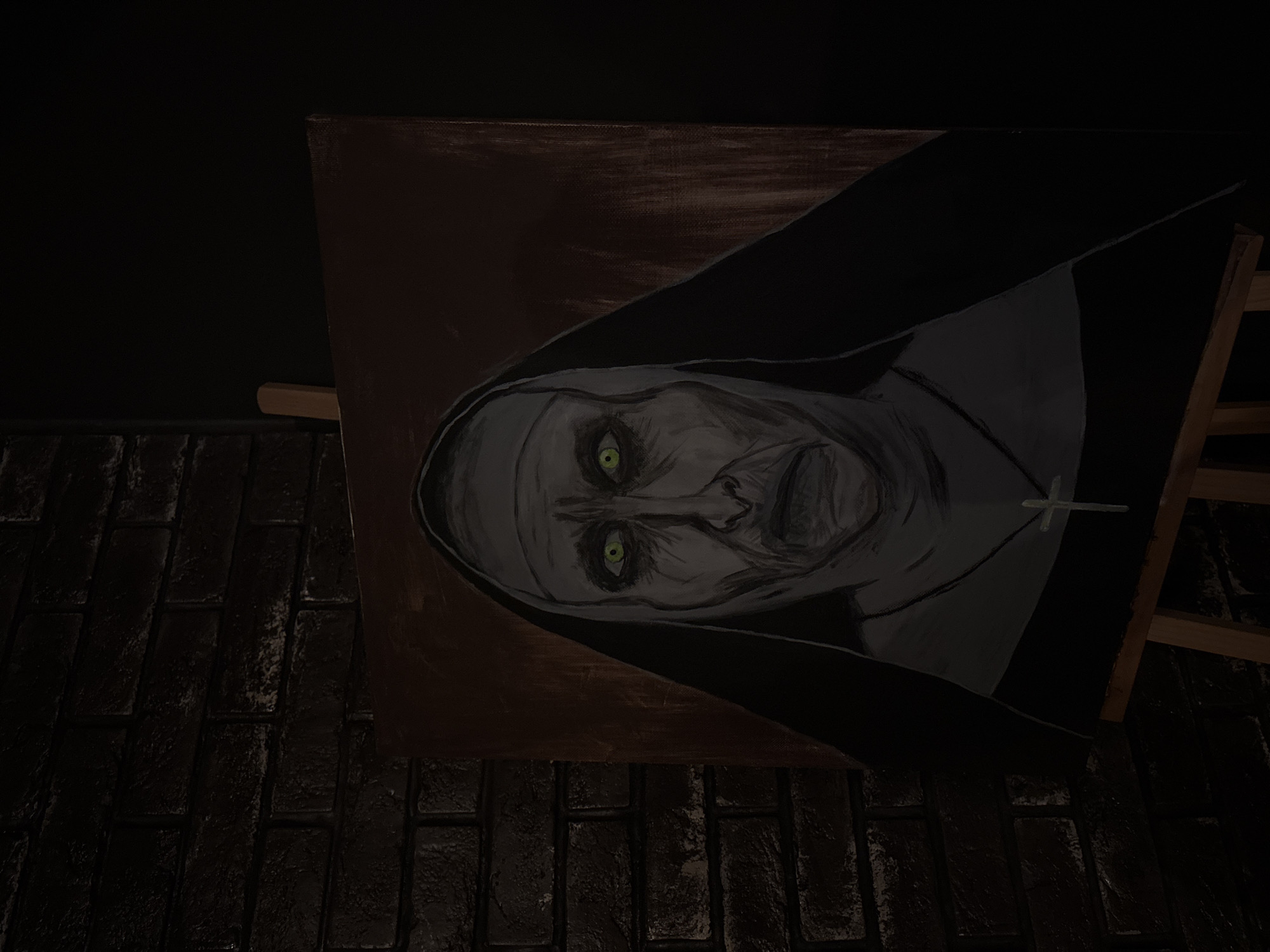 An oil painting of the demonic nun Valak from The Conjuring film series is displayed on a wooden easel against a dark brick wall. The painting depicts the nun's face with glowing green eyes, gaunt features, and a habit covering her head and shoulders. A simple cross pendant is visible around her neck. The setting is dimly lit, emphasizing the eerie subject matter. The location is identified as Sosnovo, Russia.