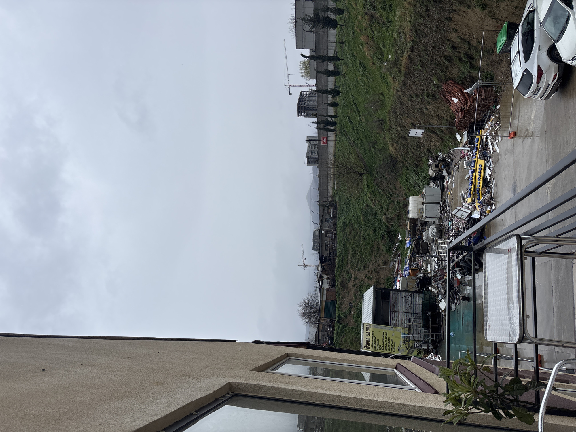 The image shows an outdoor scene, likely taken from a balcony in Tbilisi, Georgia. In the foreground, there is a building with windows and a visible balcony area with plants and a table. Beyond that, the middle ground features a cluttered area with what appears to be a large collection of miscellaneous debris and discarded items, including metal scraps, possibly automotive parts, and general refuse.
Further in the background, there's a grassy slope leading towards a series of buildings under construction, with cranes visible among them. The sky is overcast and grey. Cars are parked on the side next to the debris, suggesting the possibility of an unregulated scrap yard.