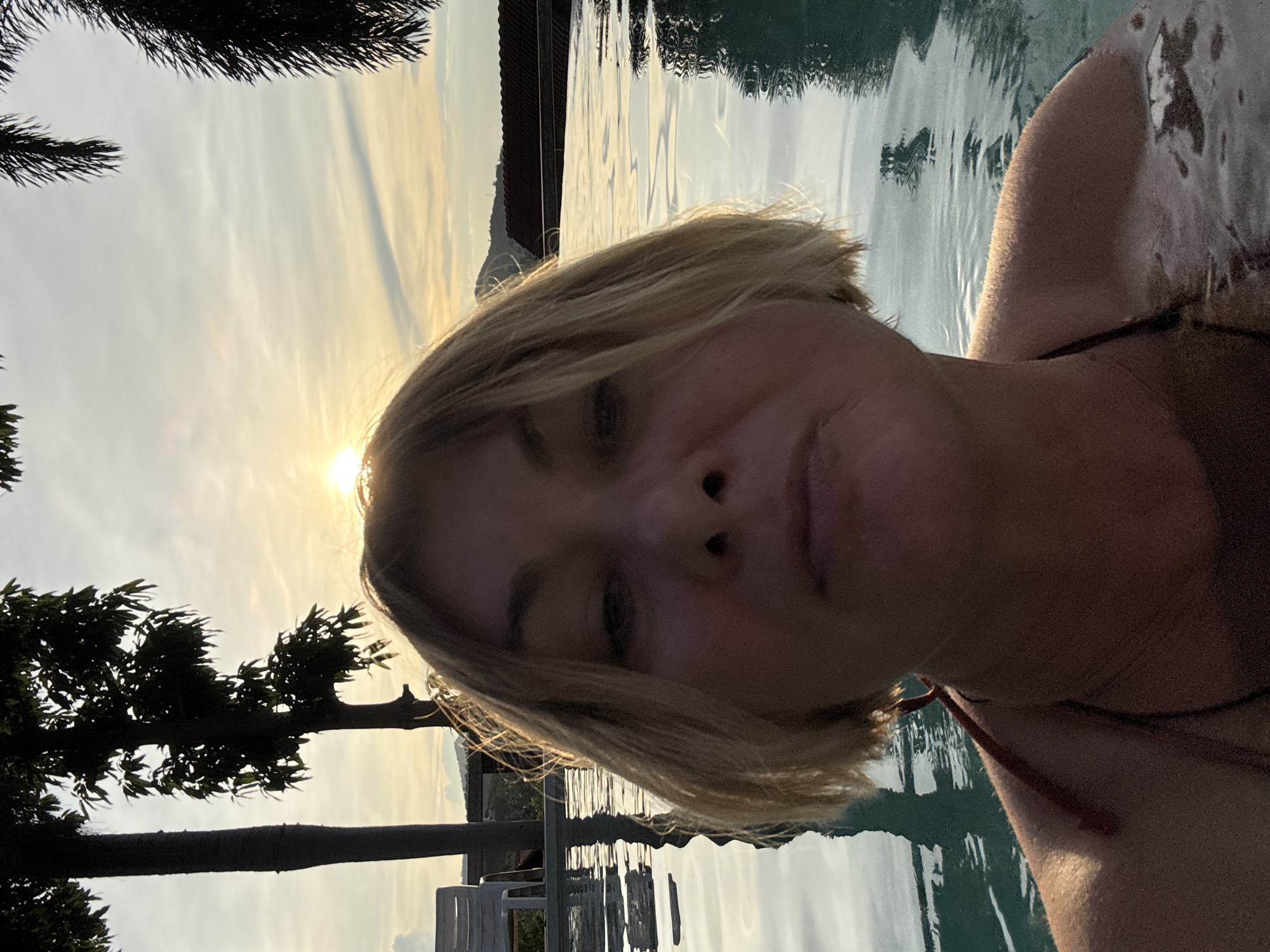 In a selfie taken from water level, a woman with short blonde hair is looking directly at the camera, her face illuminated by the setting sun. She is in a swimming pool, with ripples on the surface of the water and reflections of the sky. Palm trees and other foliage are visible in the background, silhouetted against a soft, cloudy sky with the sun low on the horizon. A white chair and a building with a tiled roof are also visible in the distance. The overall impression is a relaxed and scenic setting.
