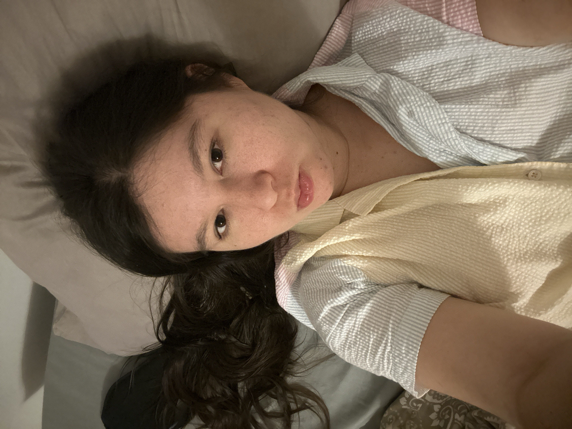 A woman with long, dark, wavy hair lies on a gray pillow and possibly a bed. She is looking directly at the camera with a neutral expression. She is wearing a light-colored, textured striped shirt or pajama top with alternating stripes of white/blue and white/yellow. The photo is taken from a slightly above and angled perspective. The lighting is soft and diffused. There is no evidence to suggest this image was taken in Câmara de Lobos, Portugal.