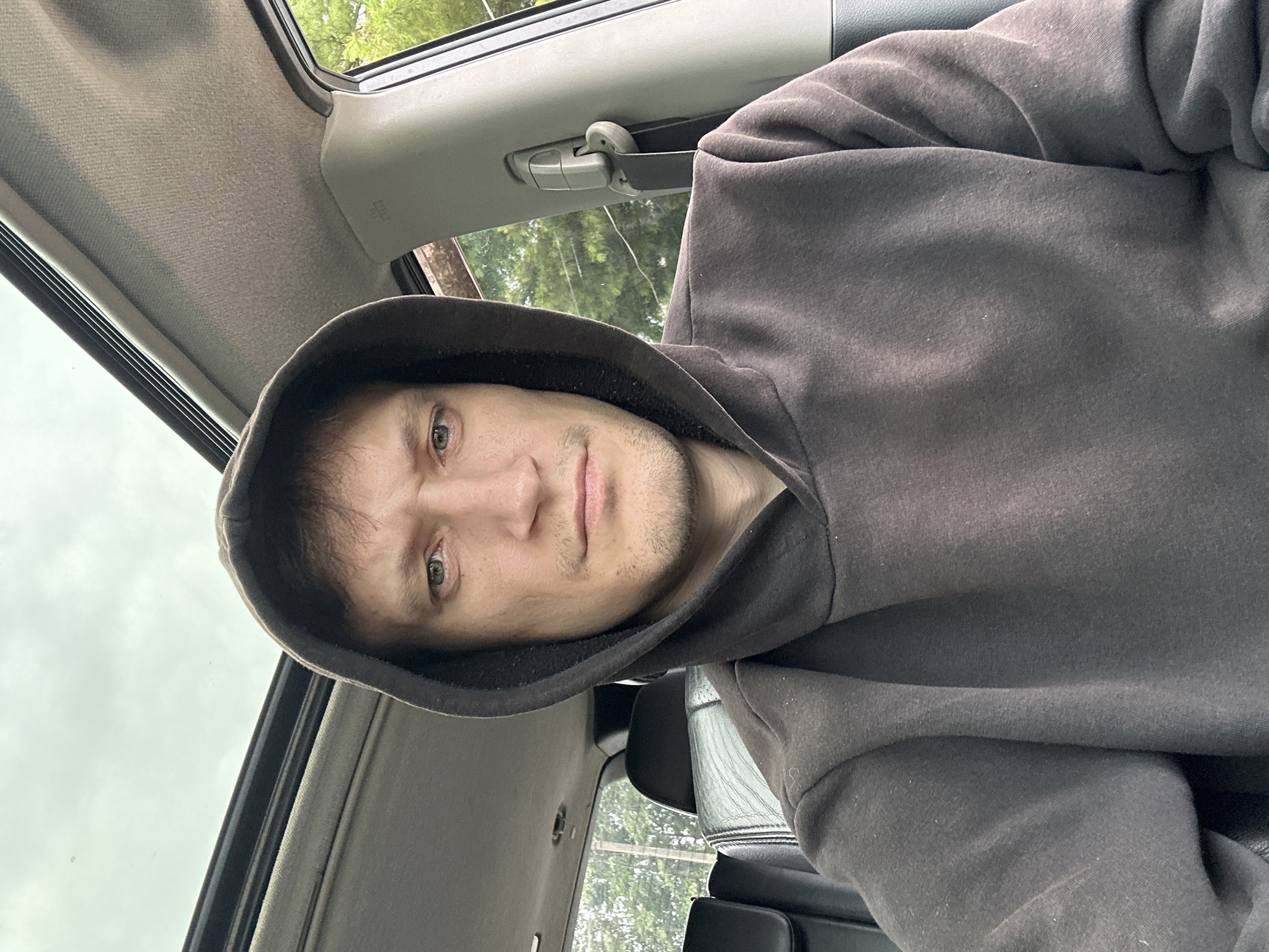 A young adult male wearing a dark grey hooded sweatshirt is seated in the driver's seat of a vehicle. The vehicle has a sunroof that is open, and the interior appears to be a light grey. The male is looking directly at the camera with a neutral expression. The background outside the car windows shows greenery, suggesting an outdoor location. The scene is well-lit, possibly due to the open sunroof and overcast sky. There are no specific landmarks visible that would indicate the location in Makiv, Ukraine.