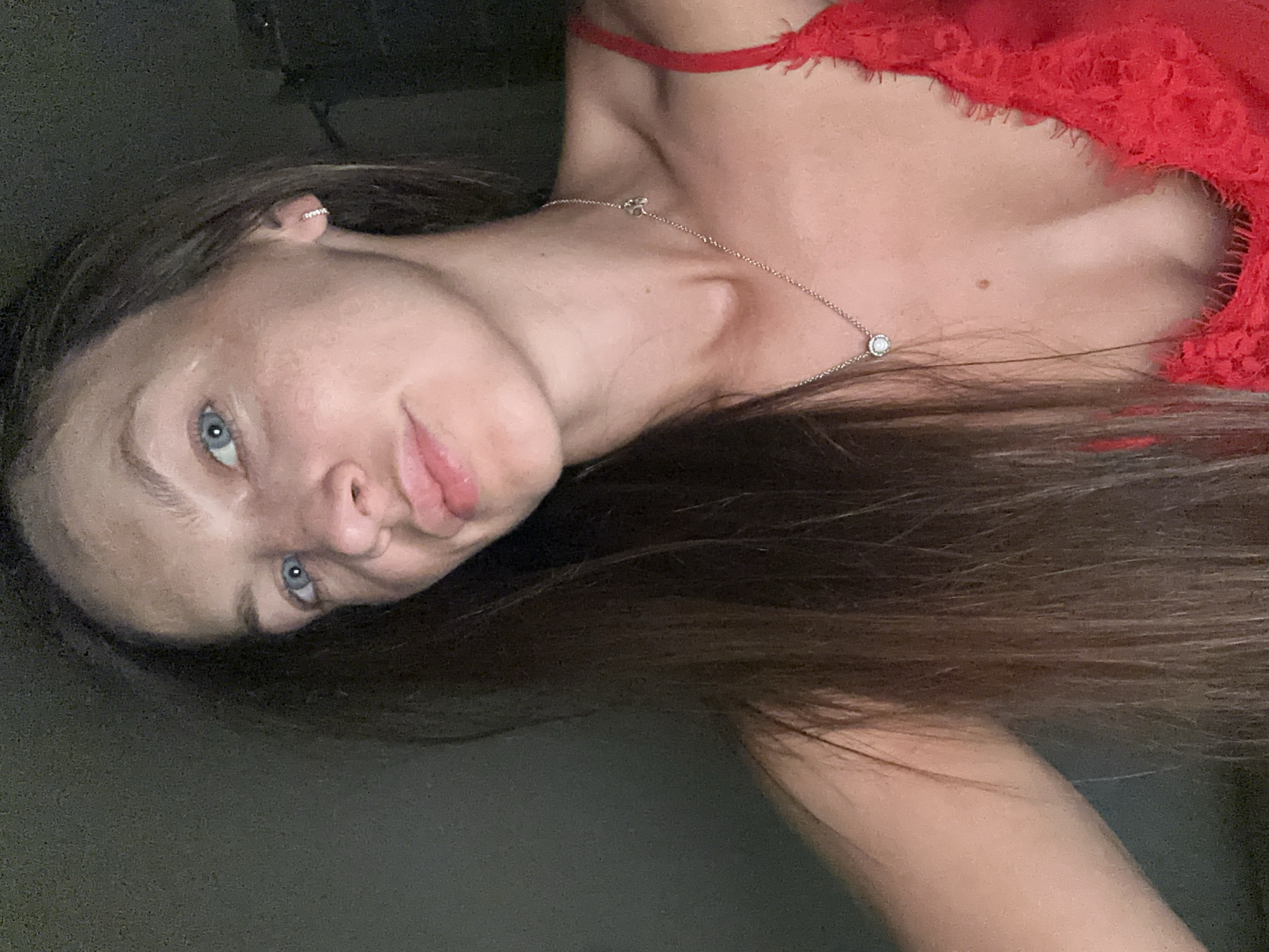 A woman with long brown hair and blue eyes is looking up and slightly to her left. She is wearing a red lace top and a silver necklace with a small circular pendant and a paw print charm. The background is dark and out of focus, suggesting an indoor setting. There is no indication of the location being in Donji Pobori, Montenegro.
