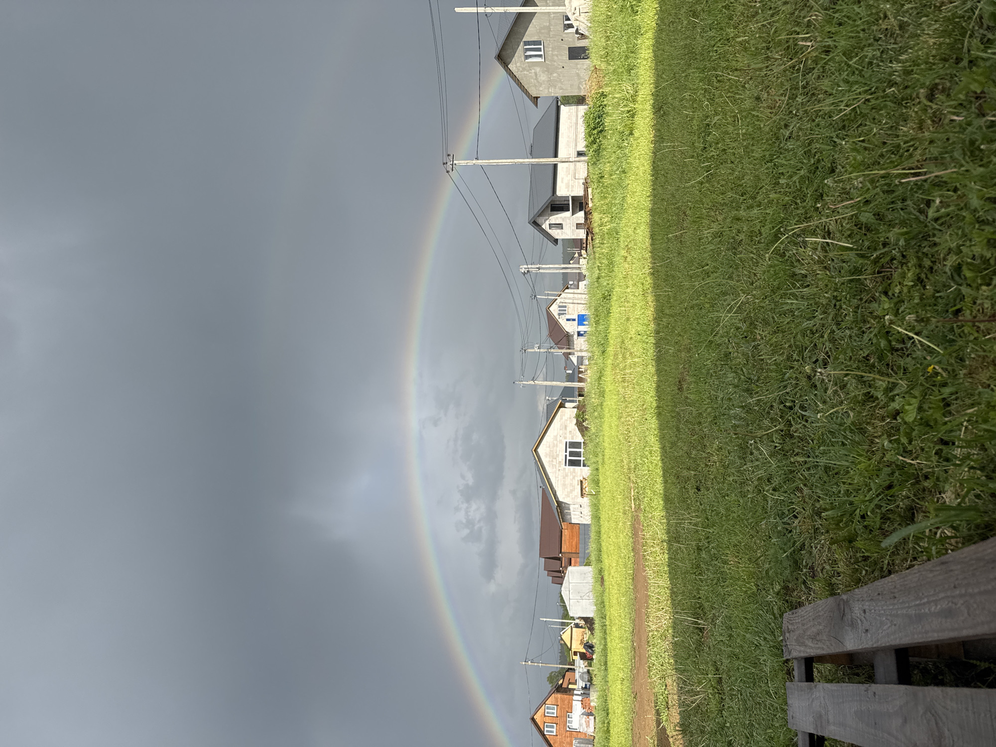 The image shows a view of a residential area in Ферма, Russia under a dark, overcast sky featuring a distinct double rainbow. The perspective is from ground level, with a wooden structure partially visible in the foreground. To the right of the image, there is green grass. Residential buildings of varying designs, including partially constructed homes, are visible lined up along the rainbow's curve. Power lines and utility poles run alongside the houses. The color palette is dominated by grays and greens, with the vibrant colors of the rainbow standing out against the dark sky.