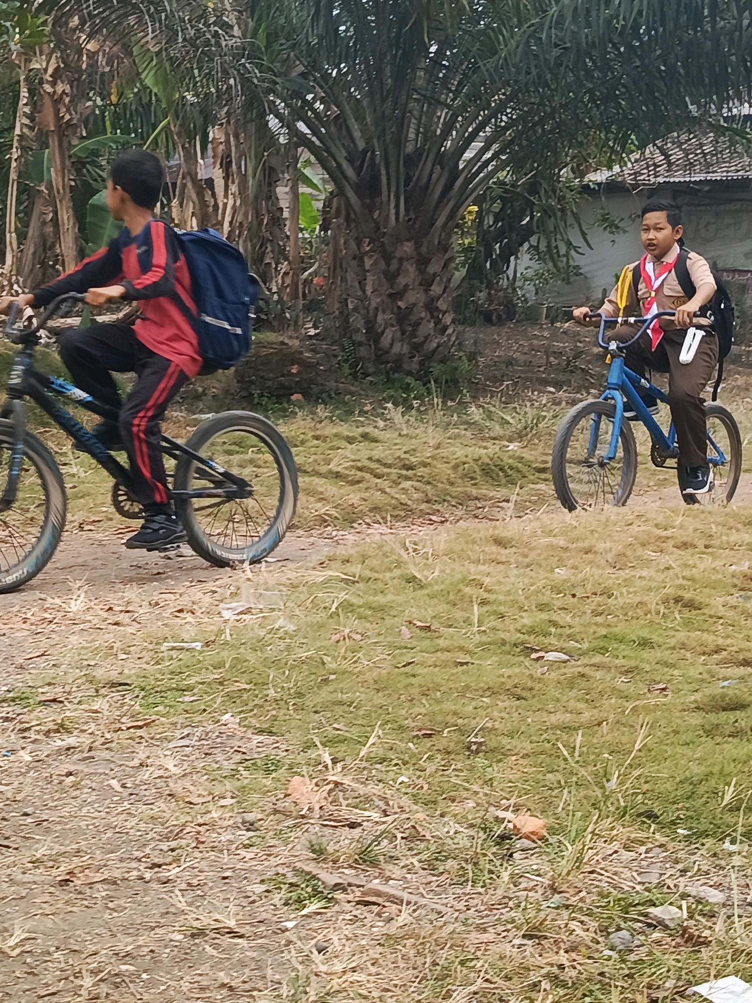 The image shows two young boys on bicycles on a dirt path in an outdoor environment. The boy on the left is wearing a red and black long-sleeved shirt, black pants with red stripes, and a dark blue backpack. He is riding a dark-colored bicycle. The boy on the right is wearing a scout uniform with a tan shirt, brown pants, a red and white scarf, and a dark backpack. He is riding a blue bicycle. Both boys appear to be in motion. The background features palm trees, other vegetation, and a structure with a tiled roof. The ground is covered with dry grass and dirt. The scene is consistent with a rural or suburban setting. This location is identified as Megang Sakti II, Indonesia.