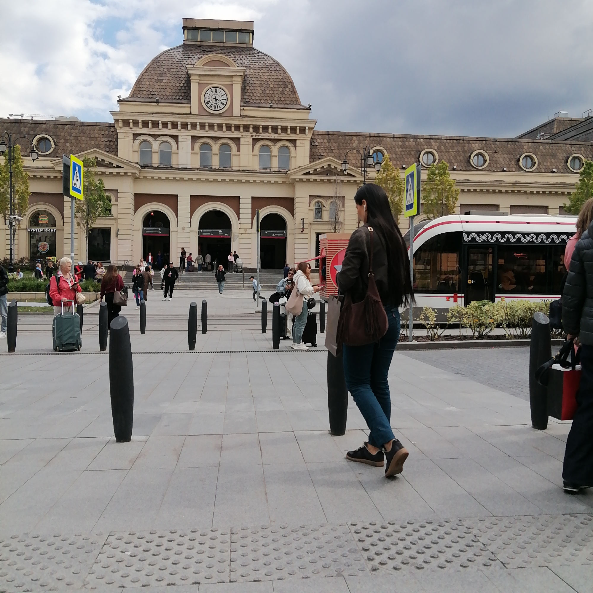 The image depicts a public space in Moscow, Russia, featuring the Belorussky railway station in the background. The architectural style is characteristic of early 20th-century railway stations, with a prominent clock tower. A tram is also visible on the right side of the frame. Several people are present, some walking toward the station entrance, and others standing around. There are black bollards. The ground is paved with gray tiles. A pedestrian crossing sign is also present.