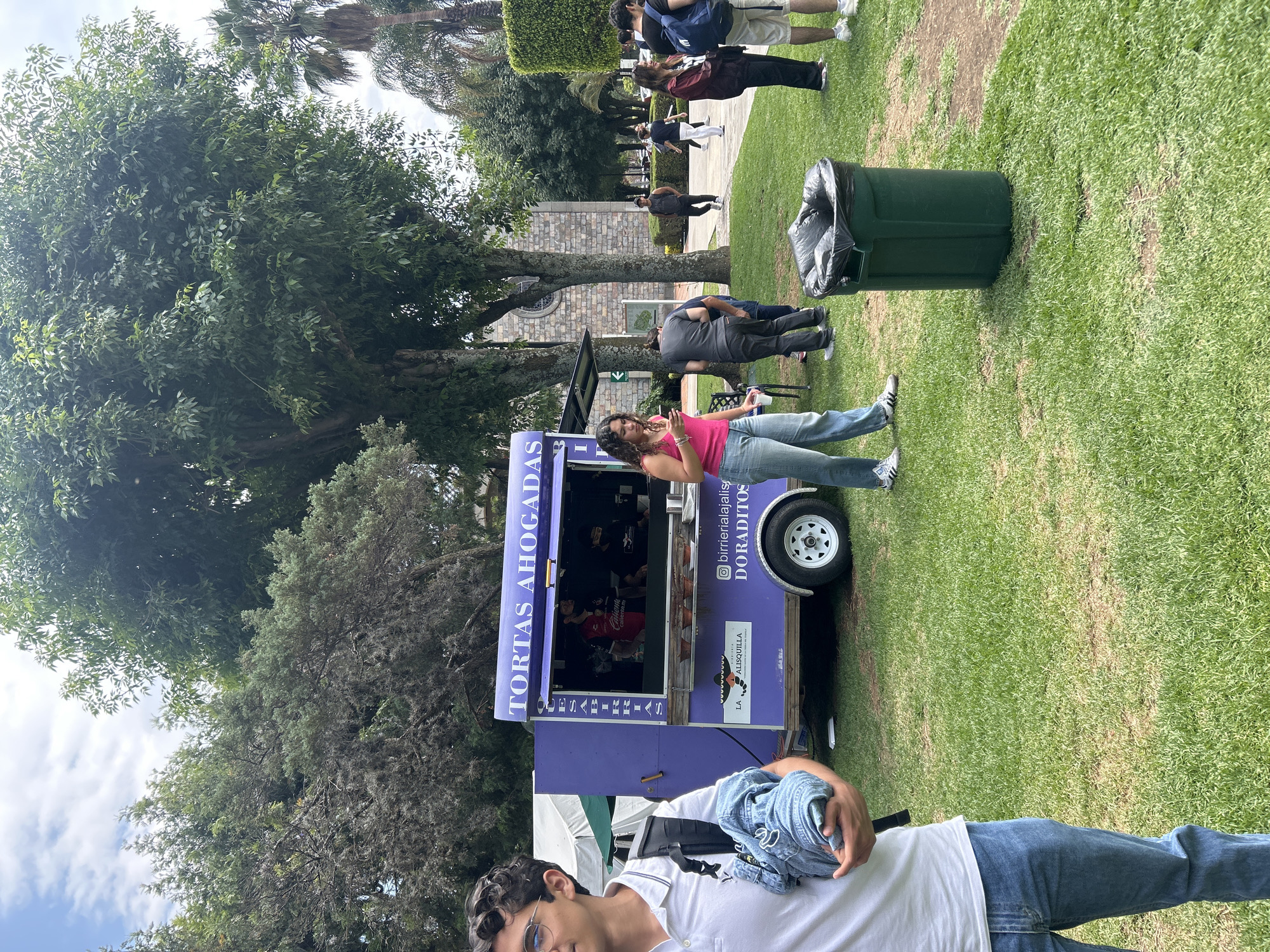 The image shows a casual outdoor scene in what appears to be a park or grassy area. A light purple food truck with the words "TORTAS AHOGADAS" prominently displayed is present. A woman in a pink top and jeans is leaning against the food truck, holding a phone in one hand and a beverage in the other. Beside her is a green trash can filled with bags.
In the foreground, partially visible, is a person in a white shirt holding a denim jacket. In the background, a group of people are walking away along a path. Trees and green foliage surround the area, and a building is visible further back. The location seems to be a public gathering place.