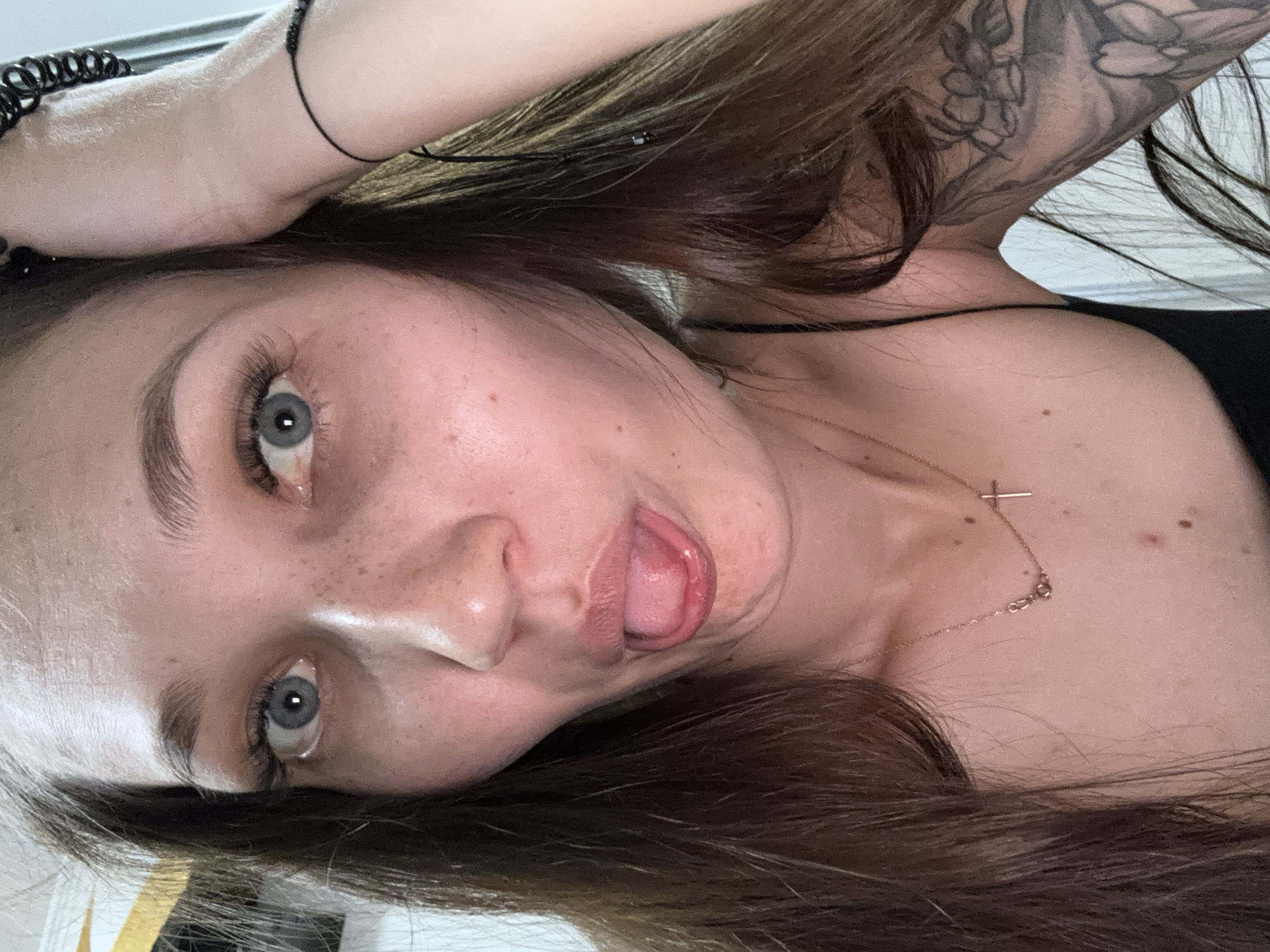 The image is a close-up selfie of a young woman with long brown hair, blue eyes, and freckles. She is sticking her tongue out and her hand is on her head. She is wearing a black camisole and a delicate gold necklace with a cross pendant. A tattoo is visible on her arm. The background is blurred but appears to be indoors with some light coming from the right. There is no visible indication of the location being Novokuznetsk, Russia.