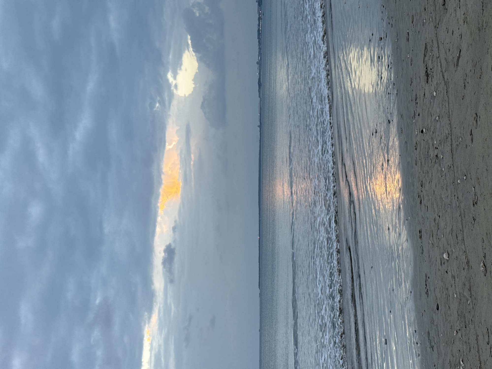 The image depicts a beach scene at dusk. The sky is filled with dramatic, swirling clouds, with a bright patch of yellow and orange light breaking through on the left side, suggesting the setting sun. The ocean stretches out to the horizon, appearing calm with small waves gently lapping at the shore. The wet sand reflects the light from the sky, creating streaks of color and texture. Small shells and pebbles are scattered across the sand, with faint footprints visible. In the distance, a faint line of lights suggests a town or city on the coast. The overall mood is serene and tranquil, with the soft light and gentle waves creating a peaceful atmosphere. The time of day is clearly evening, indicated by the low angle of the sun and the colors in the sky. The location cues of a beach, ocean, and distant lights are consistent with a coastal area. There is no visible text in the image.
